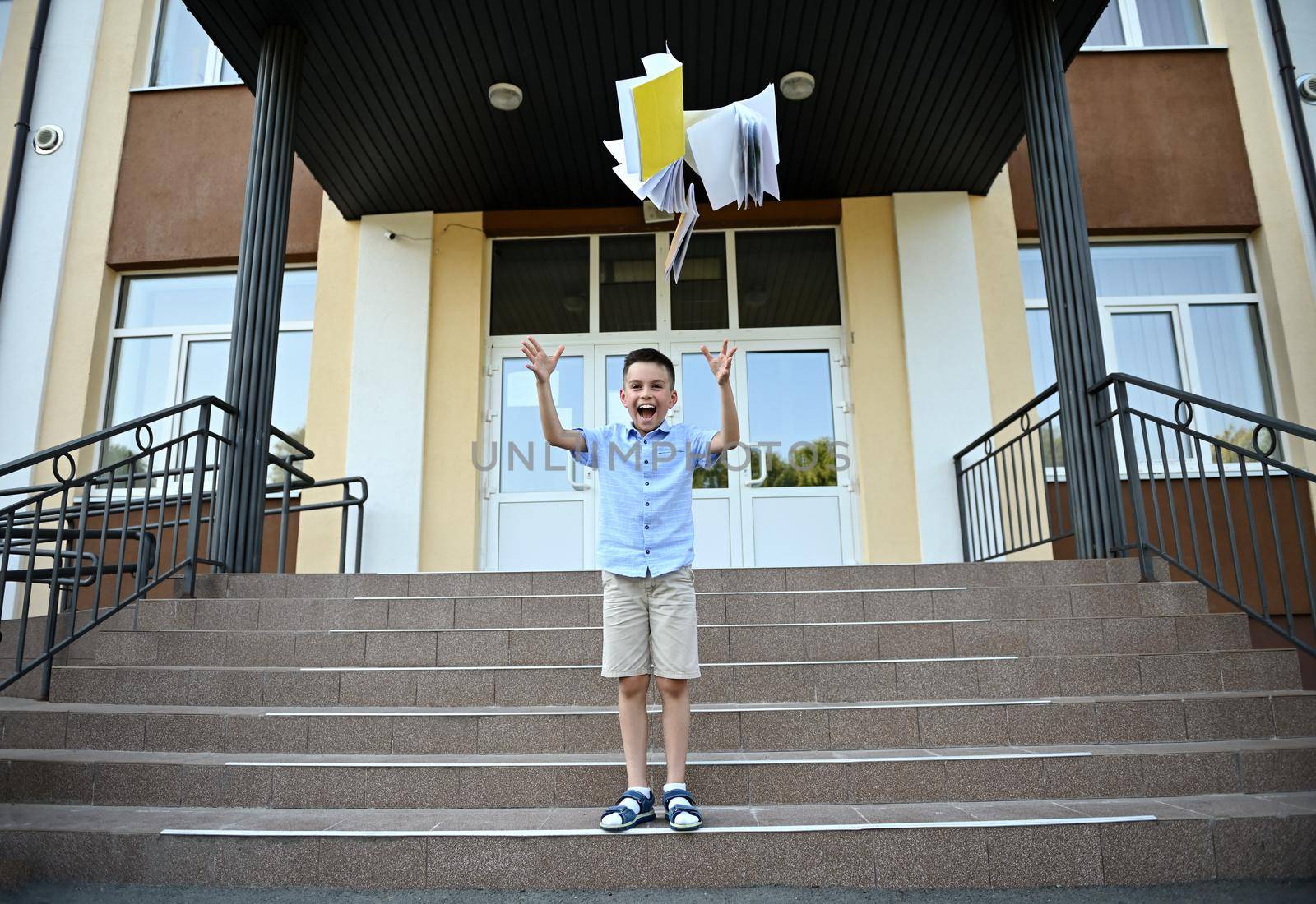 Schoolboy rejoices at the end of lessons and school, throws workbooks up, laughing and smiling happily while standing on the stairs against the background of the school institution
