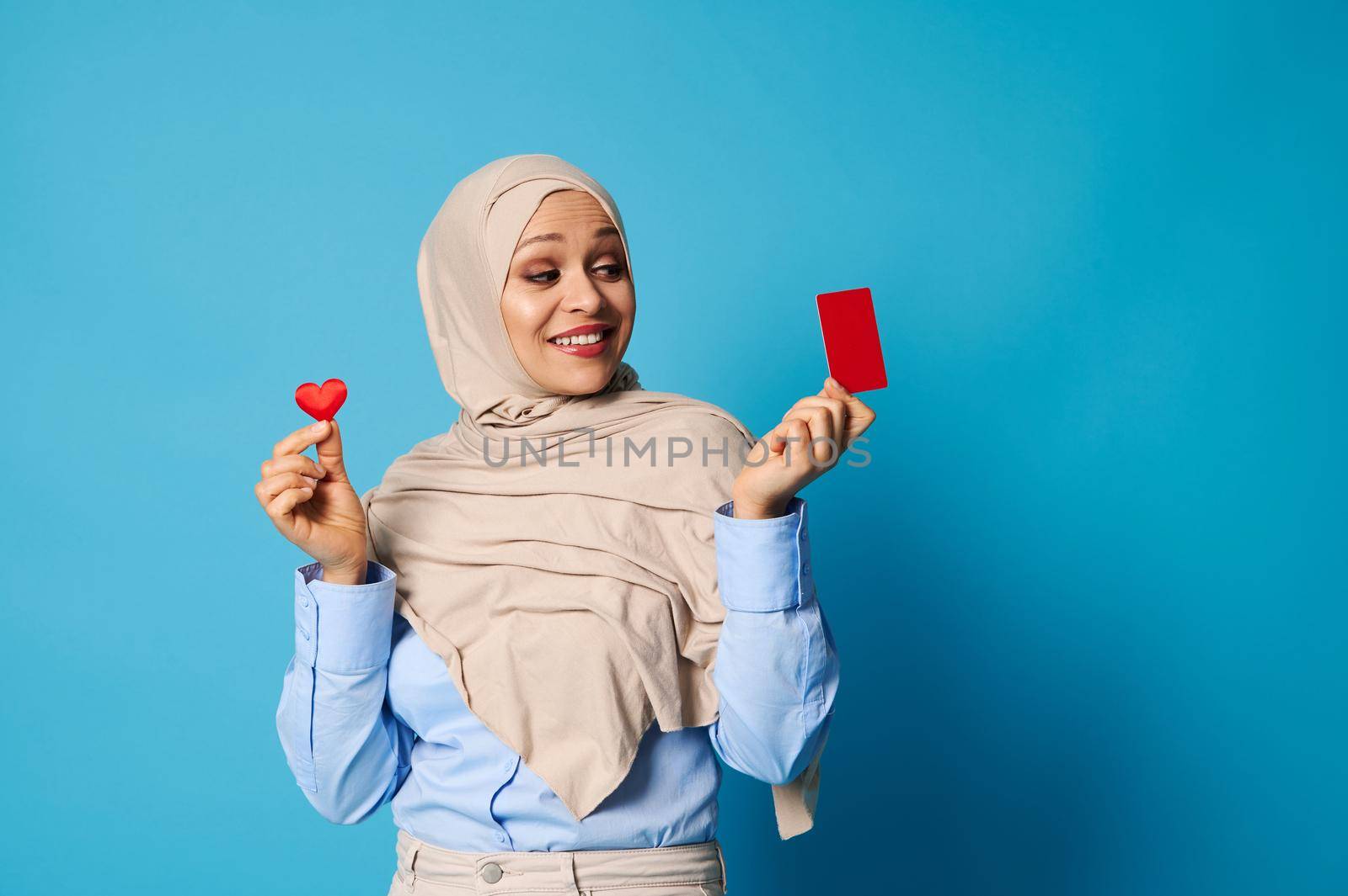 Smiling woman with oriental appearance wearing hijab, holding a shape of heart in one hand and looking at a red plastic blank card in her other hand. Blue background, copy space
