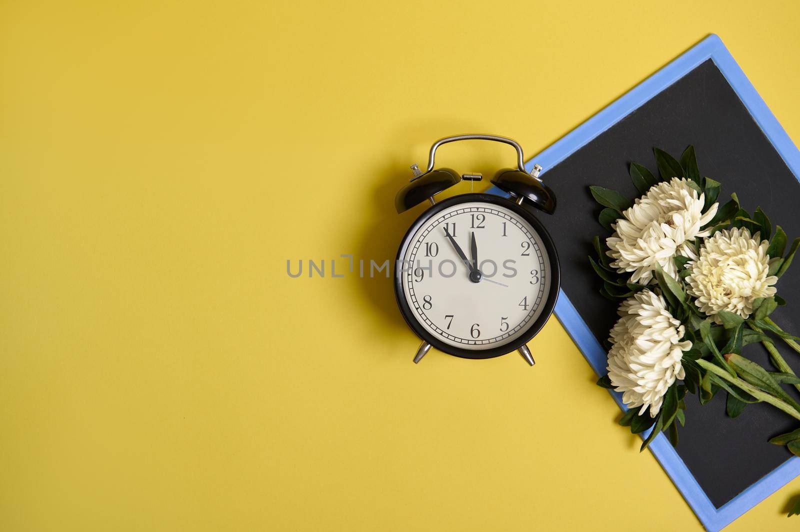 Flat lay of delicate beautiful stylish bouquet of asters flowers on a blank empty chalkboard with space for text and an alarm clock isolated on yellow background