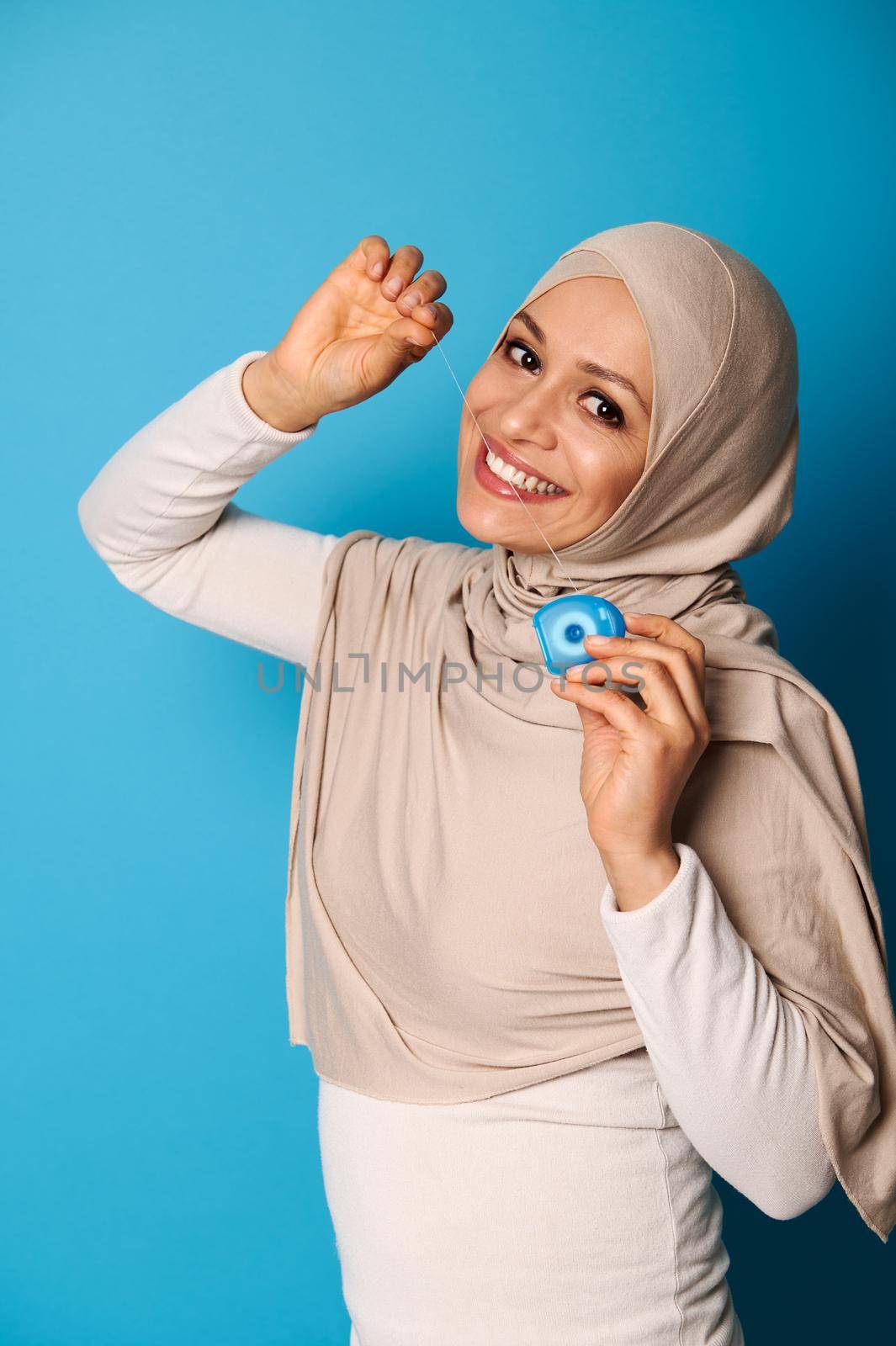 An oriental woman in a hijab smiles a toothy smile at the camera and uses a dental floss, posing against a blue background with copy space. Oral hygiene