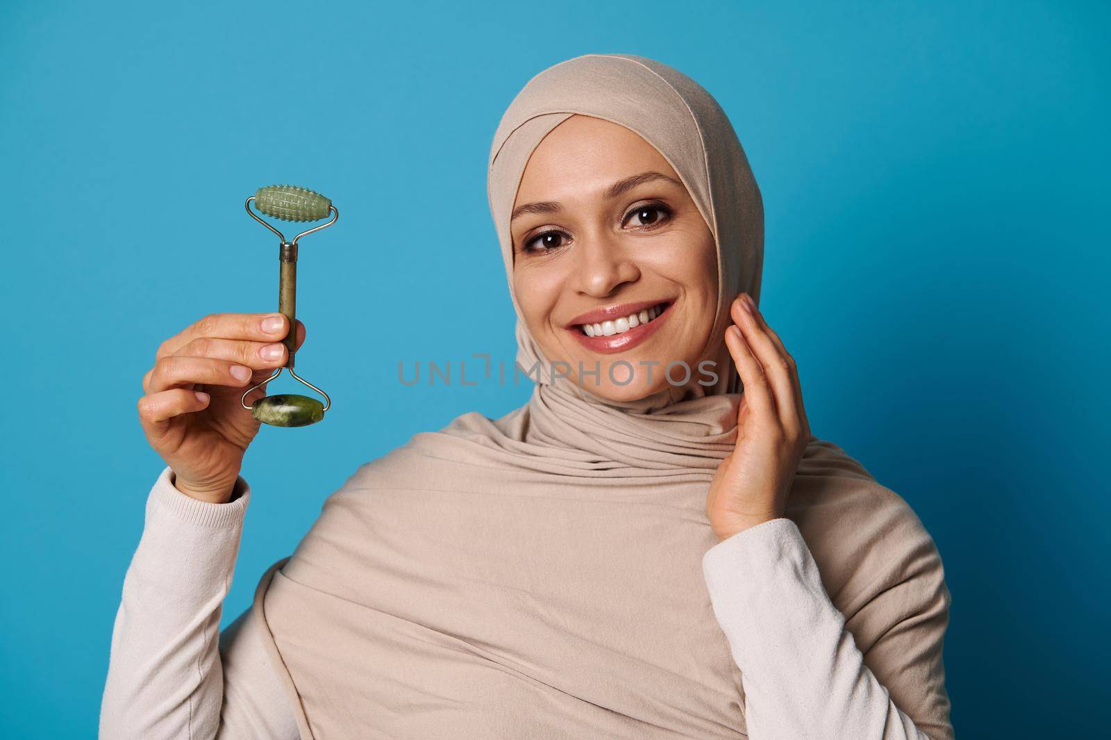 Smiling Muslim woman wearing a beige hijab holding a jade roller massager, posing looking at camera over blue background with space for text