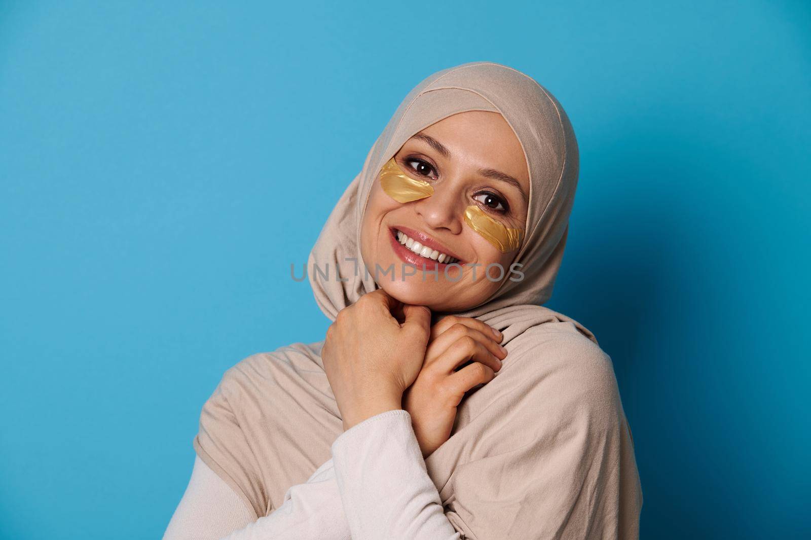 Beautiful woman with covered head in hijab, smiling at camera with hydrogel collagen patches under eyes. Isolated on blue background with copy space