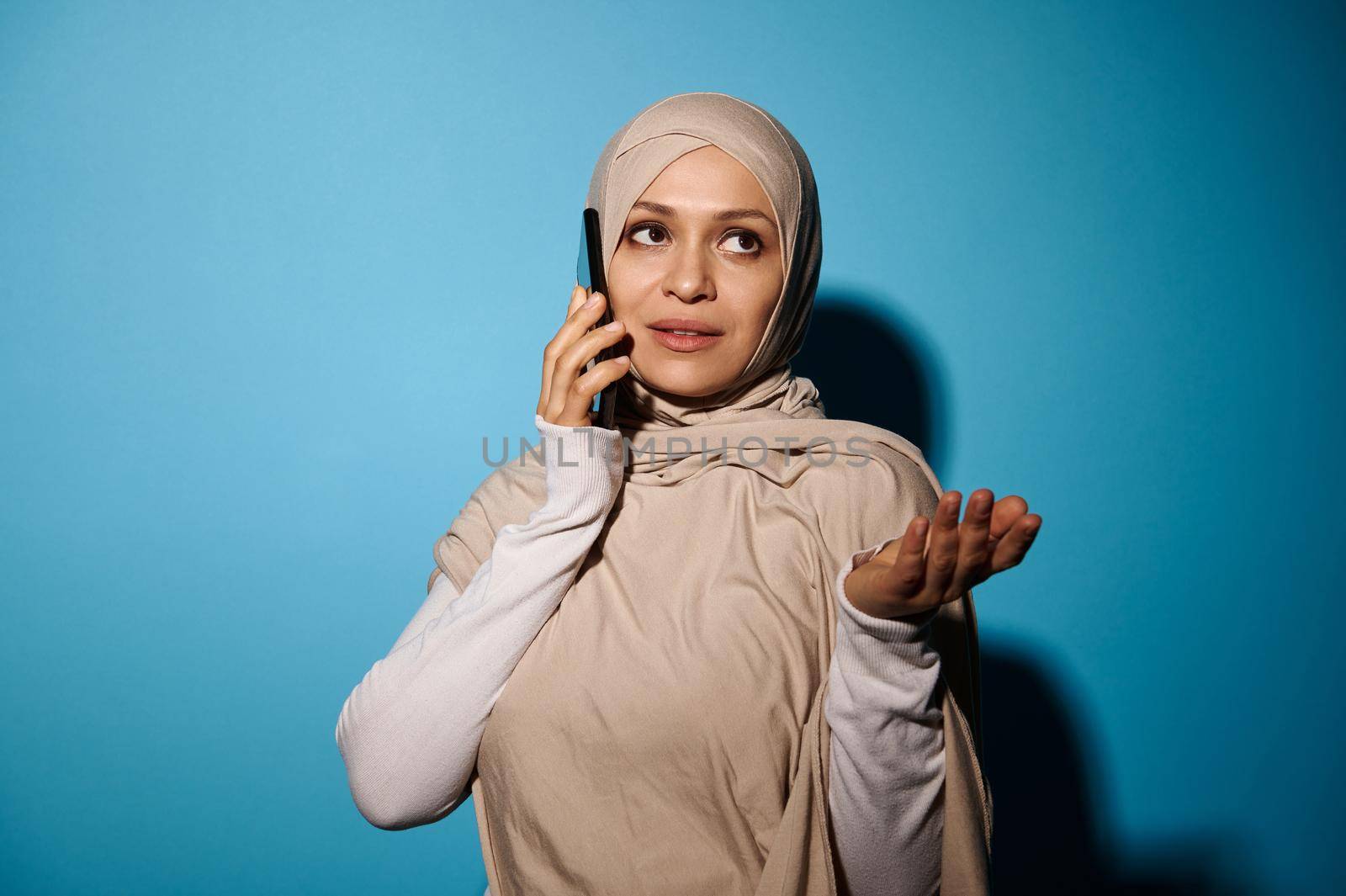 Arab Woman with covered head talking on mobile phone, and gesturing with her hand. Blue Background with copy space