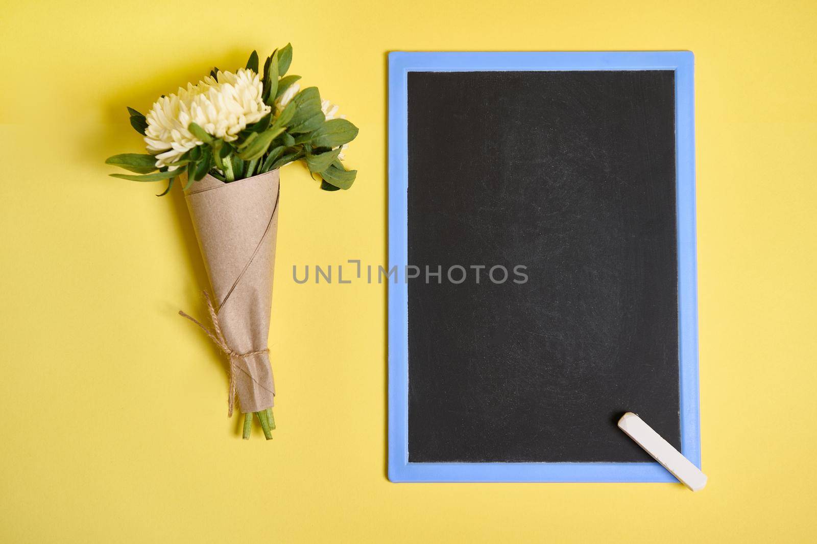 Flat lay composition of delicate beautiful stylish bouquet of asters flowers in craft wrapping paper tied with a rope and blank empty chalkboard with space for text isolated on yellow background