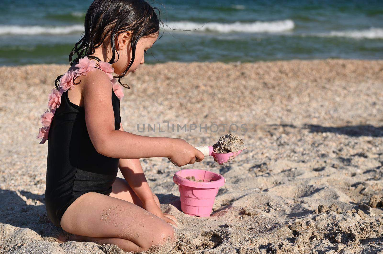 Side portrait of preschooler girl holding a rake and filling the pink toy bucket with sand, building sandy shapes and castles, enjoying sunbathing and summer vacations. Happy childhood, wellness and recreation concepts