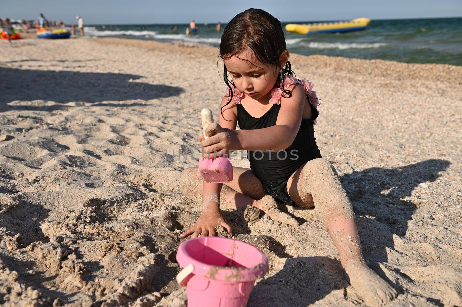 Active summer holidays concept. Baby girl playing with plastic toy bucket and shovel sitting on the sand, having sunbathing on the sea background