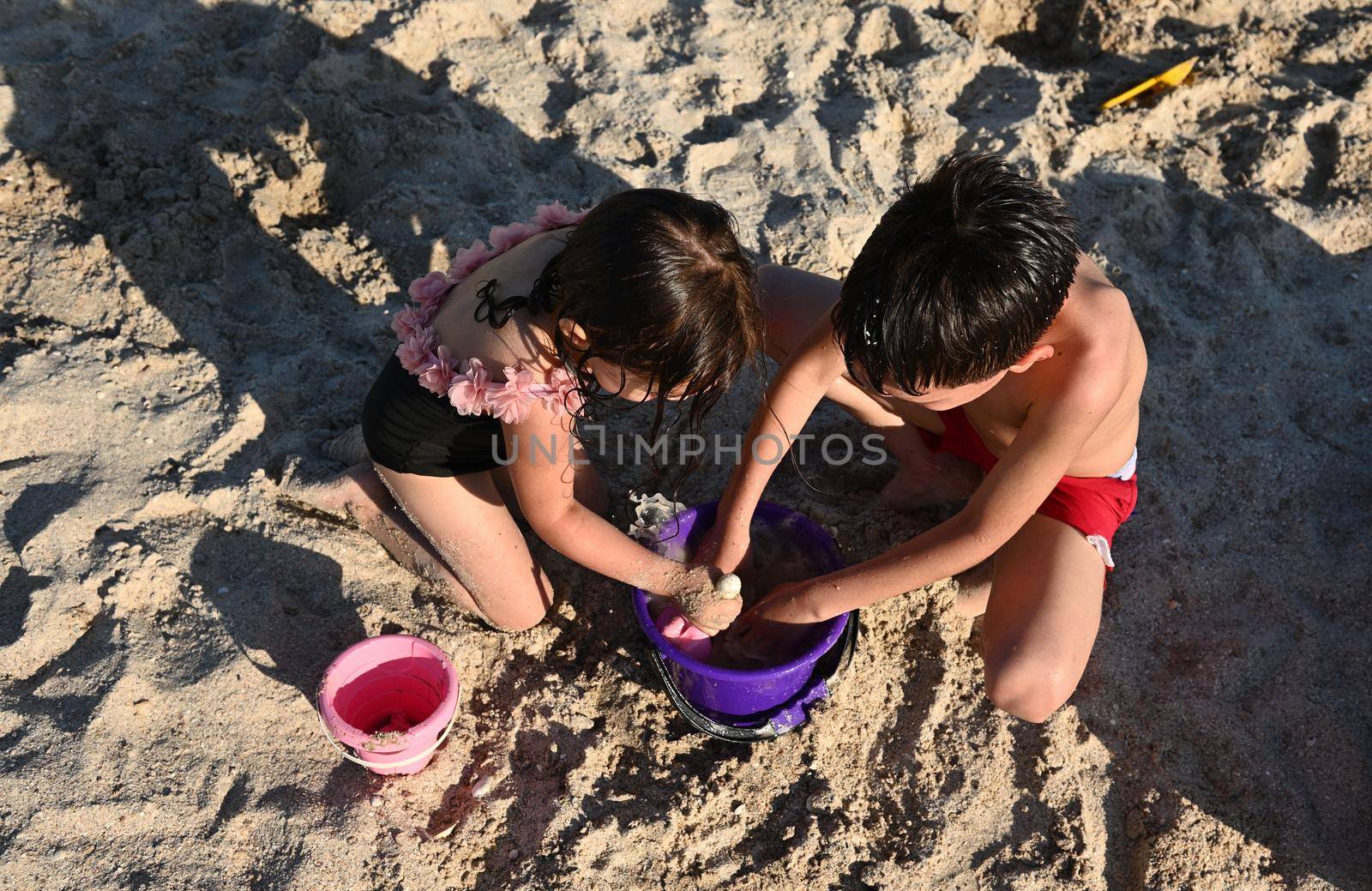 High angle view of children enjoying the construction of sand figures on the beach. Boy and a girl fill a toy plastic bucket with wet sand to build sand castles. Brother and sister having fun during summer vacation