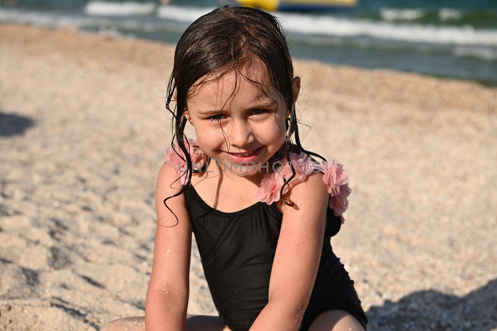 Headshot. Cute girl enjoying summer vacations. Adorable pretty baby girl with wet hair and swimsuit, sitting on the sandy beach on the sea background. Kids emotions, happiness. Summer holiday concept.