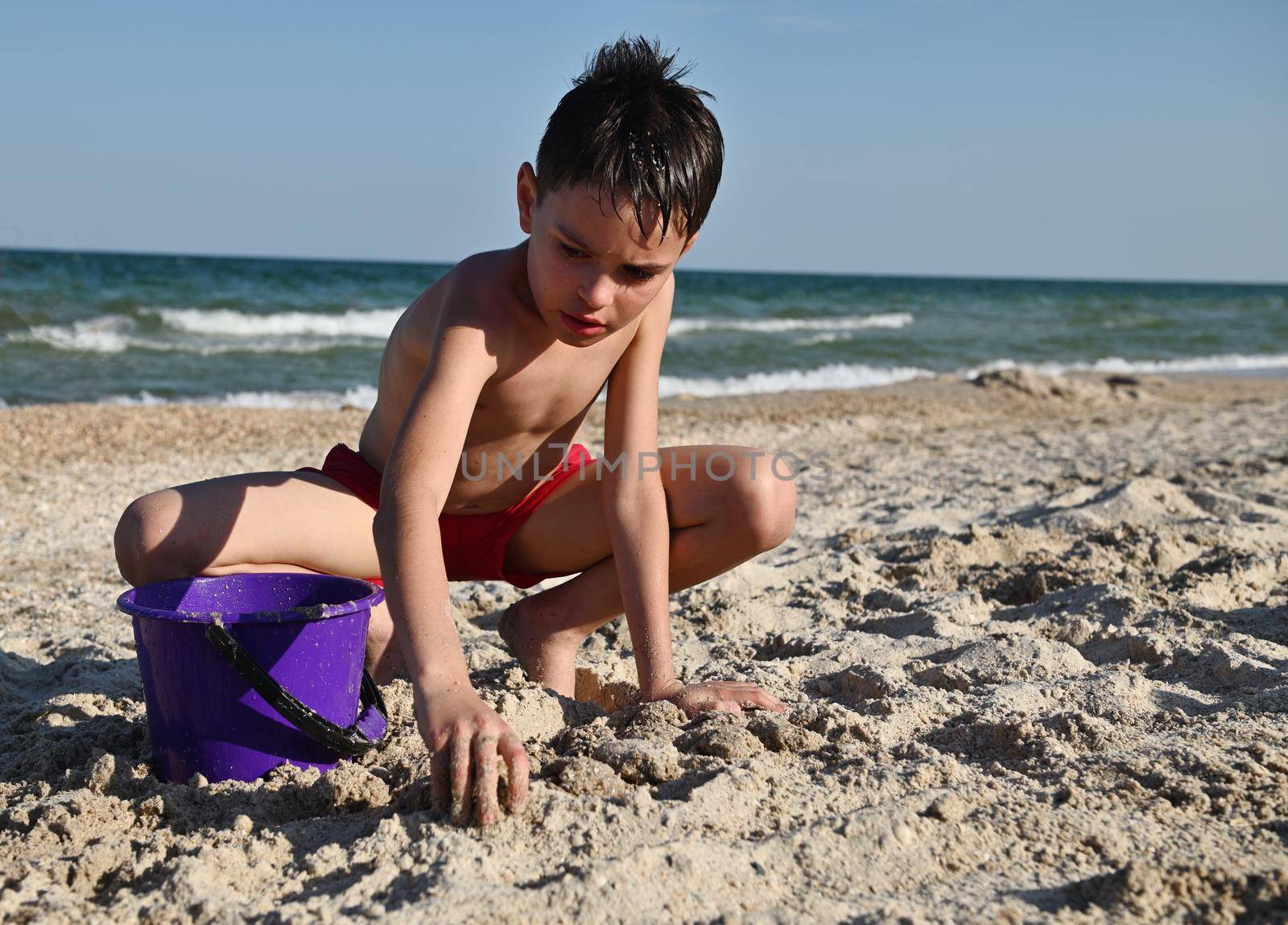 Handsome teenager in red swimming trunks playing on the sandy beach. Adorable male kid builds sand figures with wet sand against the background of the sea. Summer holidays concept. Active lifestyles and happy summer vacations