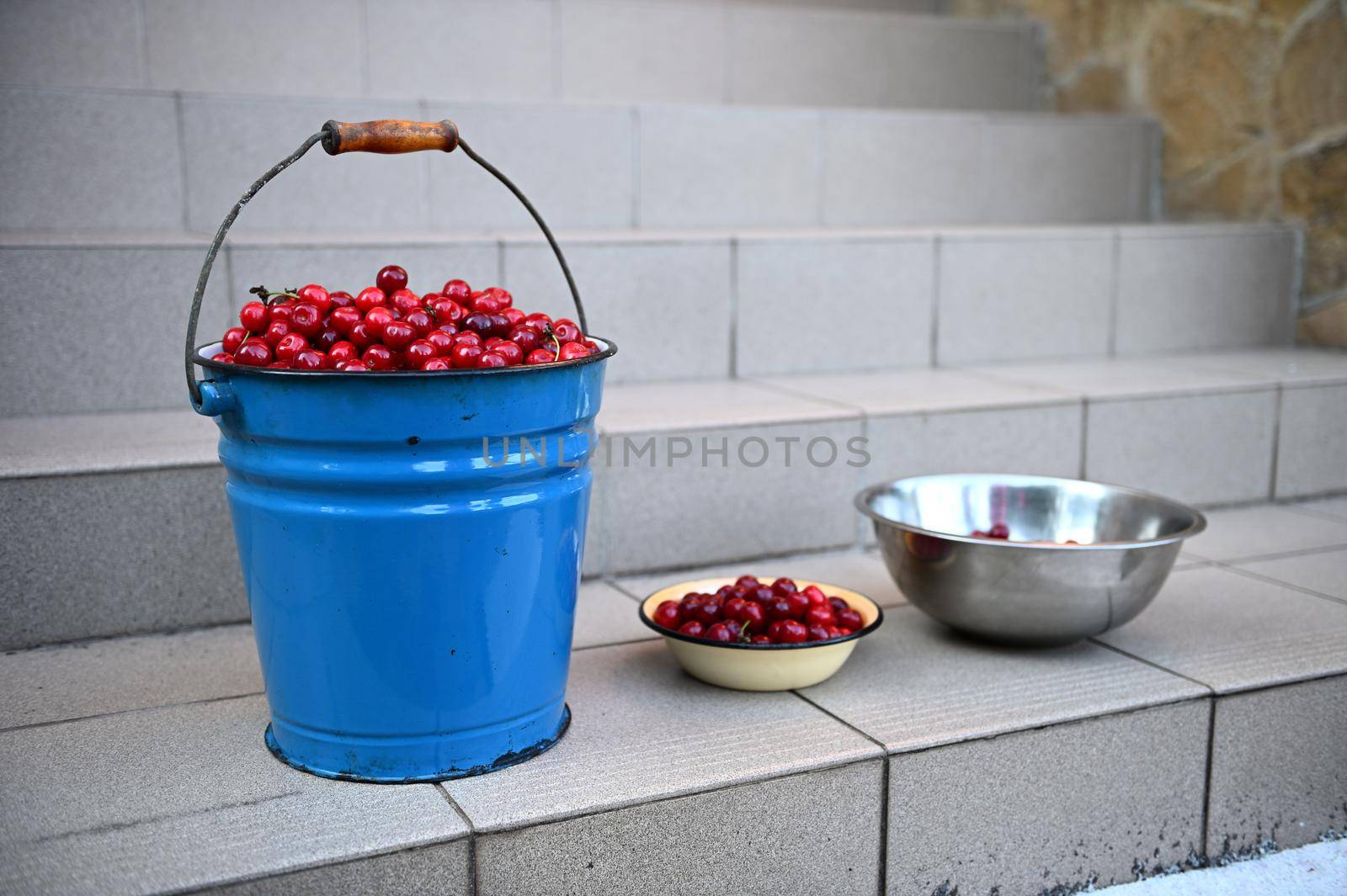 Close-up of metal bucket and bowls full of cherry harvest, standing on stairs