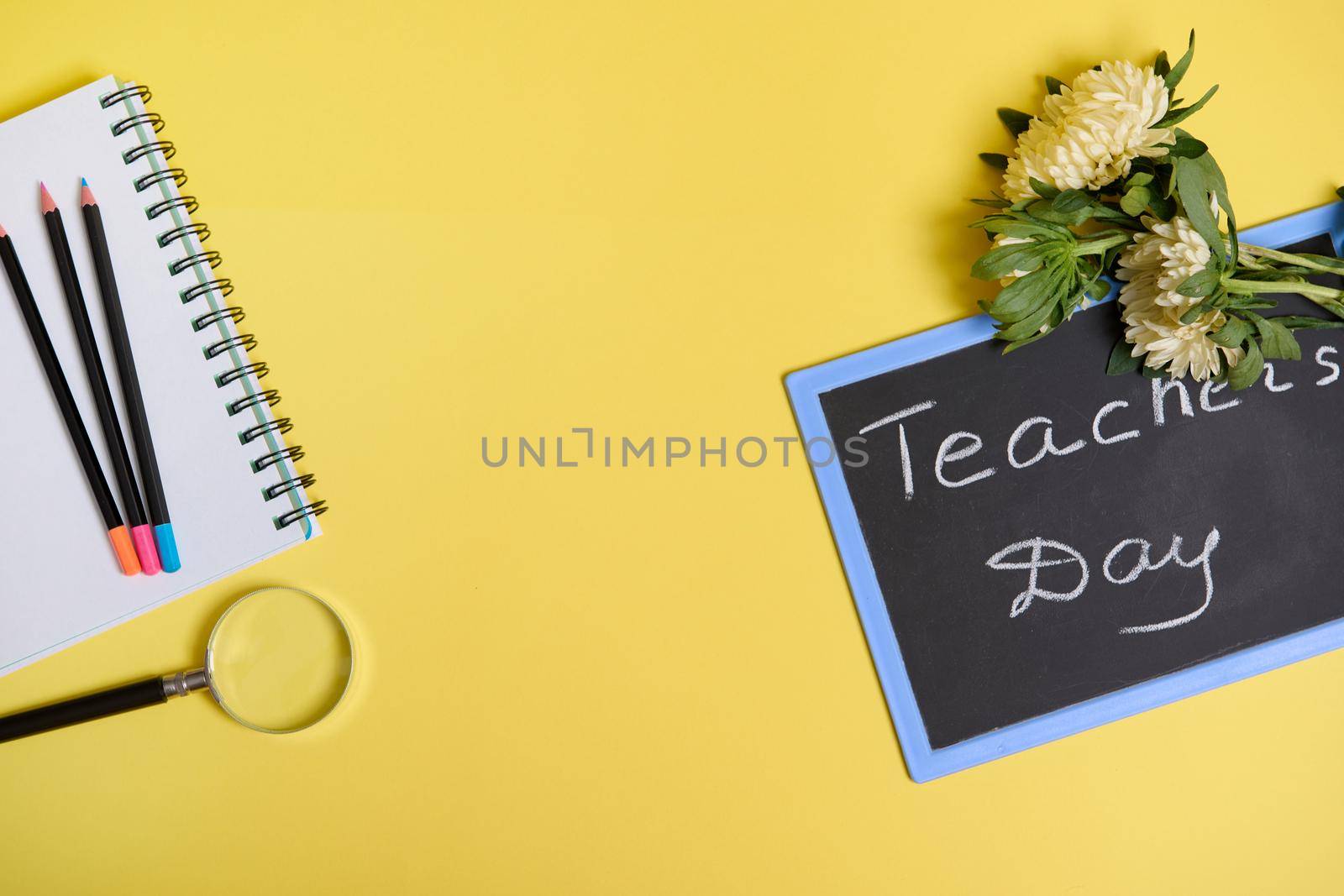 Asters flowers lying down on a chalkboard with text Teachers Day , and a magnifier loupe next to colored pencils on a blank sheets of a notepad isolated on yellow background with copy space. Flat lay