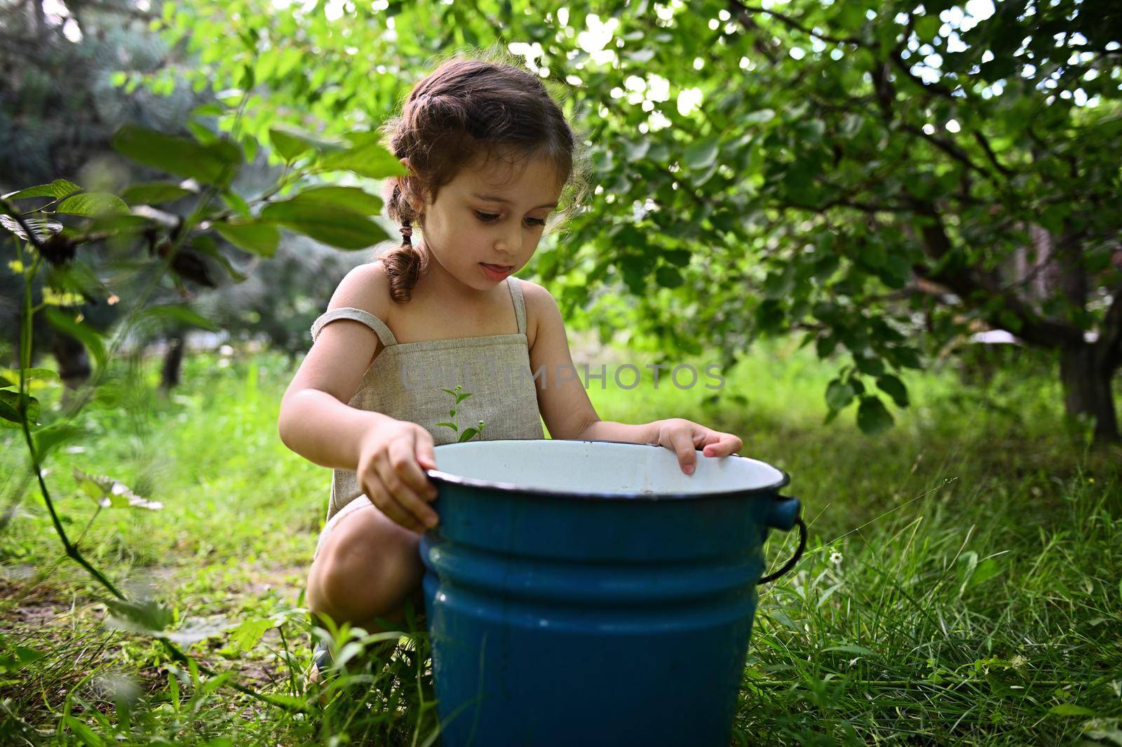 Cute baby girl sitting in the garden and peeking into a metal bucket. Gardening
