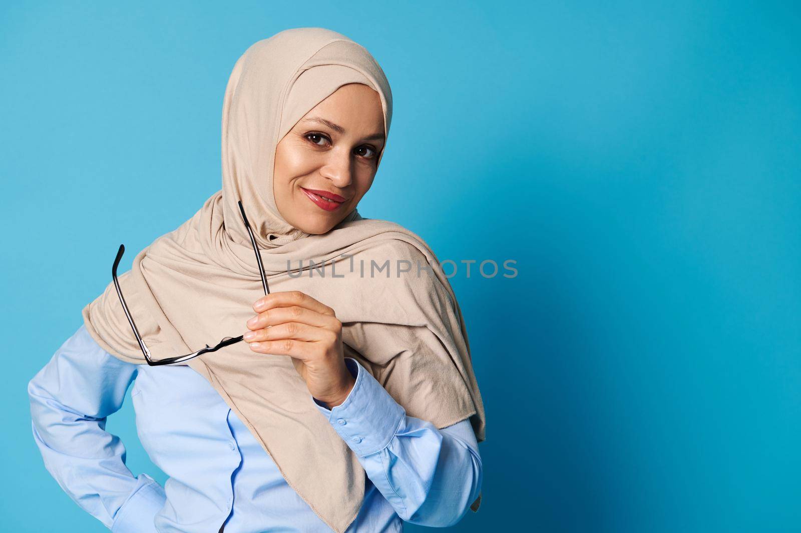 Confident portrait of an attractive Arab woman with covered head in hijab and wearing casual clothes on blue background with copy space