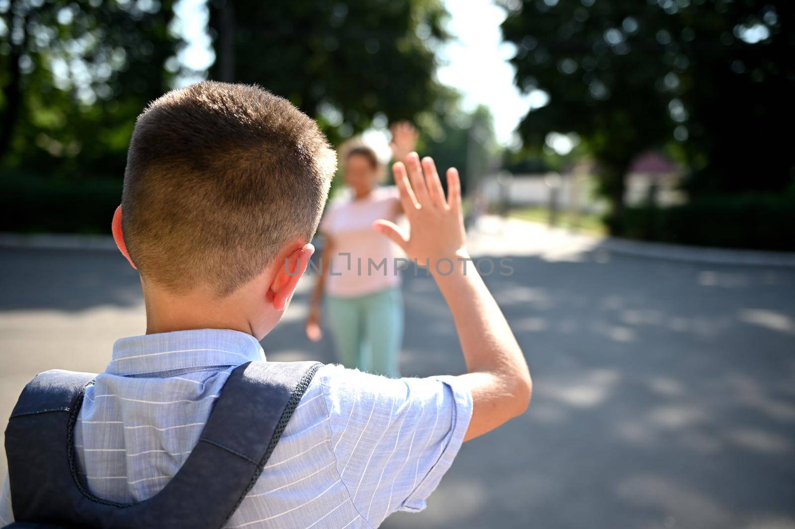 A schoolboy stands at the entrance to the school and waves to his blurred mother. The out of focus mother takes her son to school. The beginning of the school year.