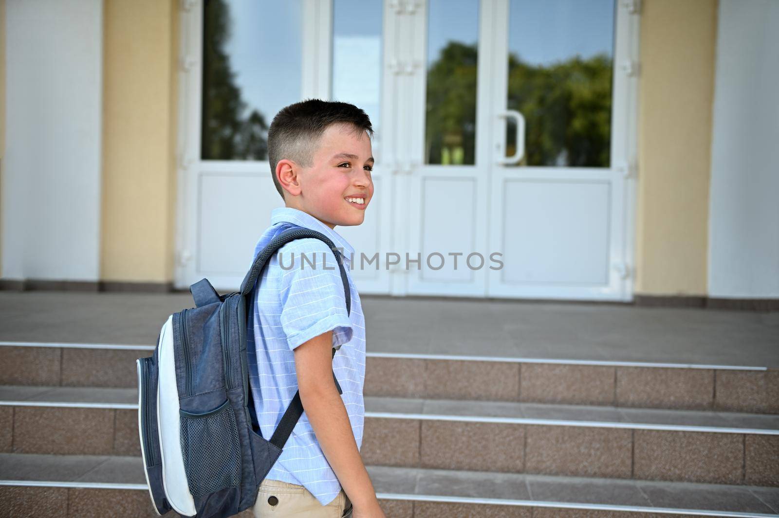 Handsome primary school student standing on the stairs in front of a school building and looking away. Copy space. Child coming back to school