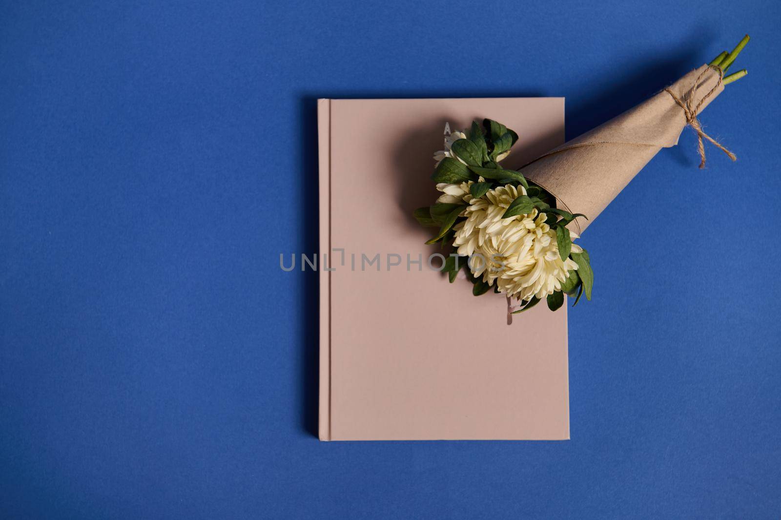 Flat lay composition of a beautiful bouquet of asters in craft paper on a beige notebook, lying on a blue background with space for text. Back to School and Teachers' Day Concepts by artgf