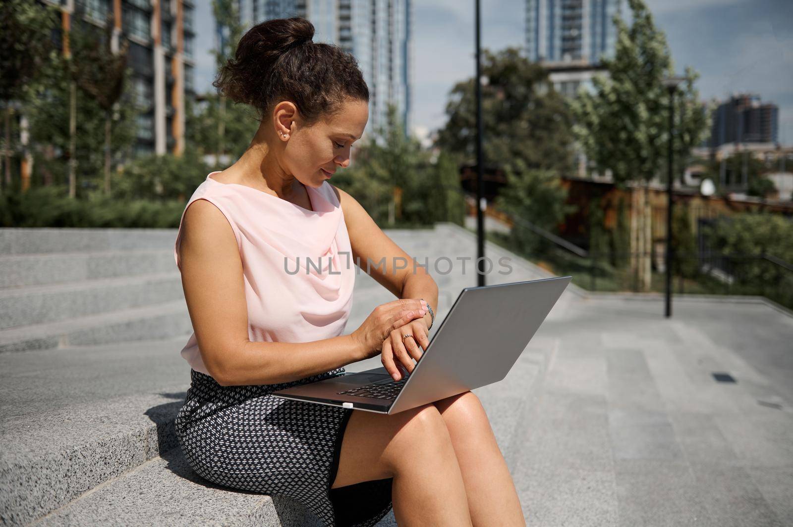 African American business woman, office worker checking time on her hand watch, using laptop while sitting on steps during coffee break or recreation. Business concept