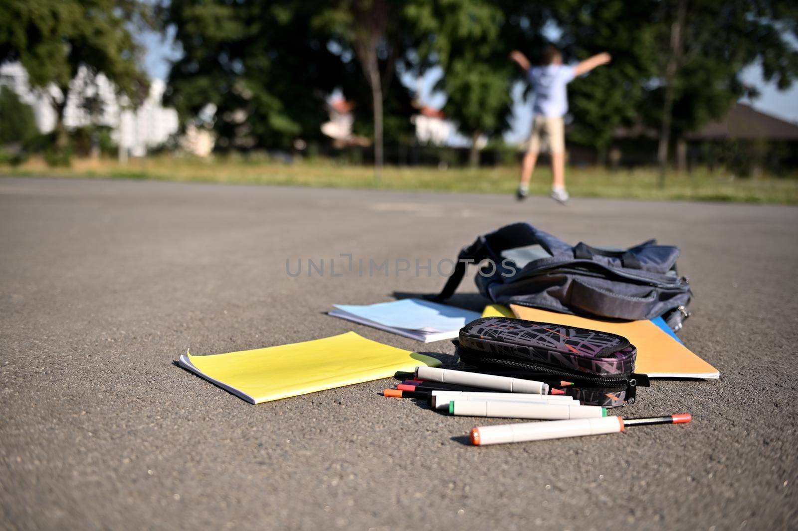 Scattered school supplies and workbooks falling out of an open backpack, lie on the asphalt of the schoolyard against the background of a blurred happy schoolboy raising his hands up by artgf