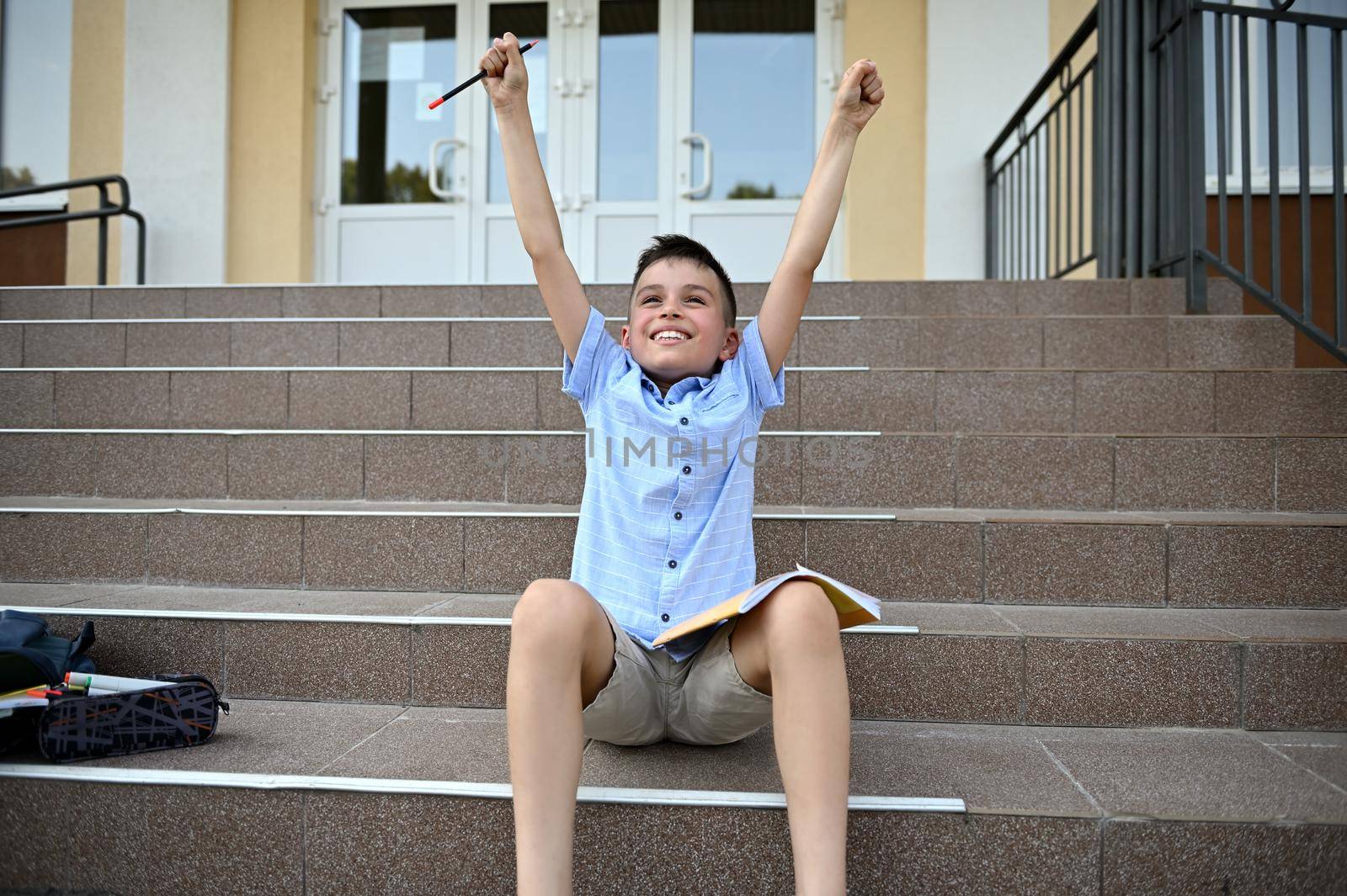 Happy elementary student raising his arms up with pencil in hand, feels happiness and satisfaction after achievement homework. Back to school concept