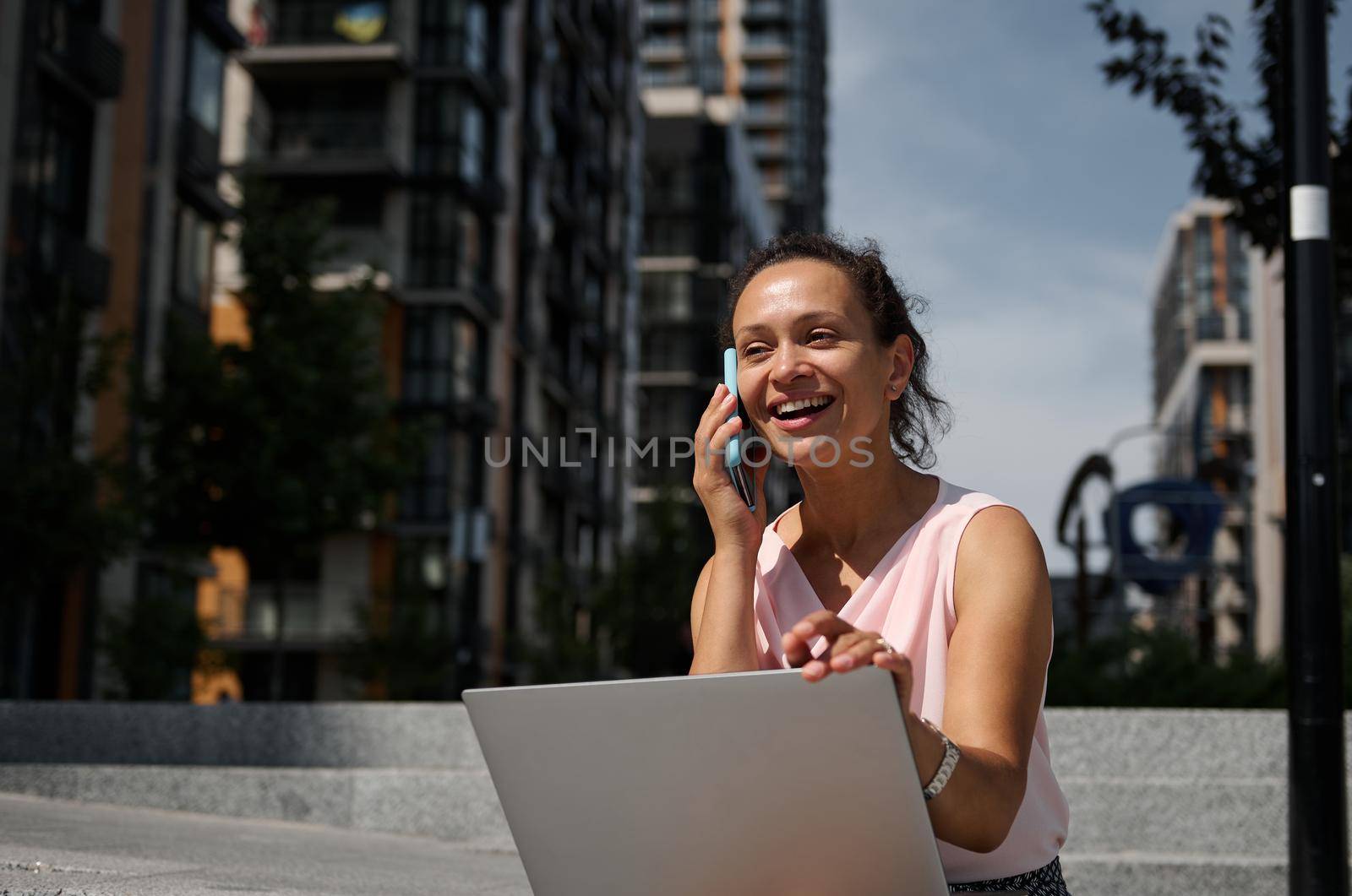 Adorable mixed race Hispanic woman talking on mobile phone, sitting on the steps with a laptop on the urban high buildings background. Business woman at lunch break time