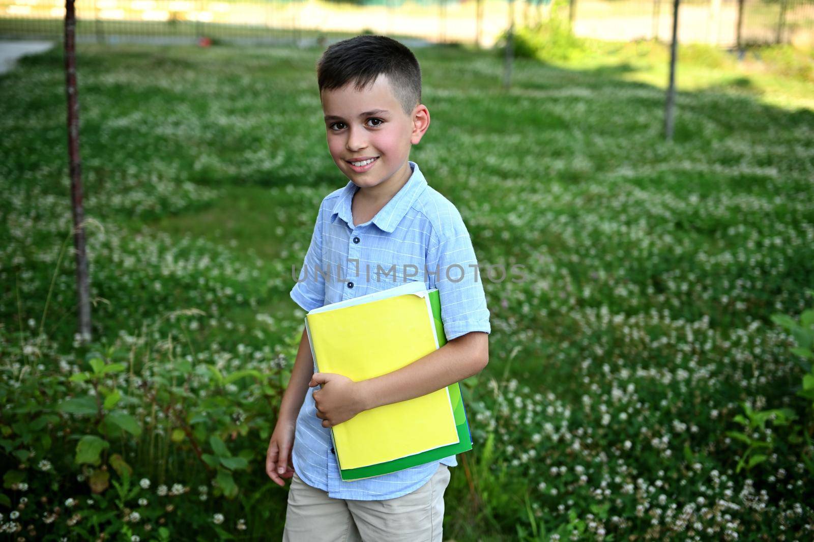 Happy elementary student coming back to school. Adorable schoolboy holding a workbooks and smiling to camera standing on green grass background.