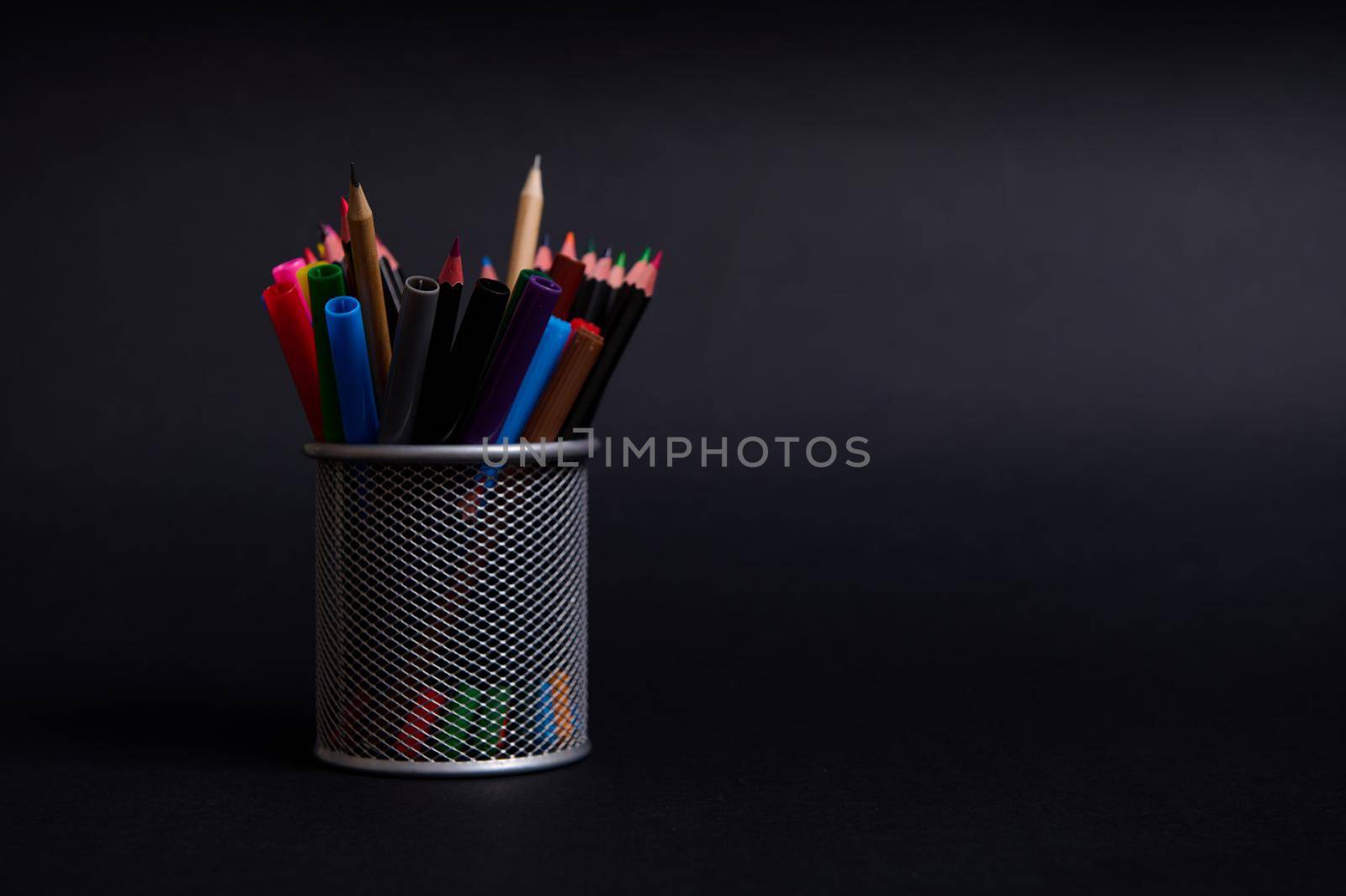Stand for pens and pencils on the desktop on a black background with copy space. Close-up of a metal bucket with stationery. Holder with colored school supplies by artgf