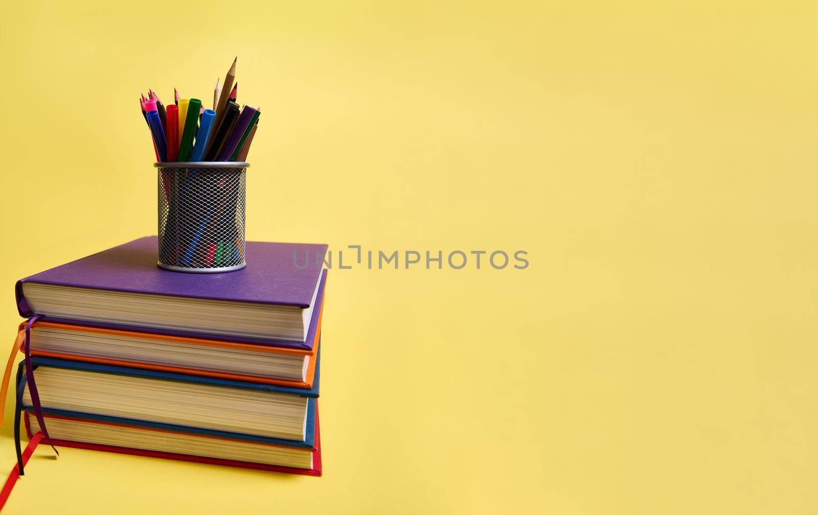 A metal pencil holder with school supplies on stacks of multicolored books. Teacher's Day concept, literary, knowledge, Back to School, education. Yellow background copy space by artgf