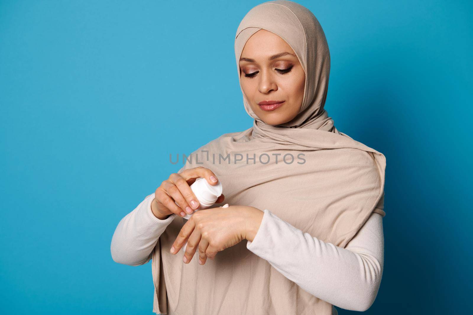 Portrait of beautiful Arab Muslim woman in hijab squeezing cream on hand, isolated on blue background with space for text