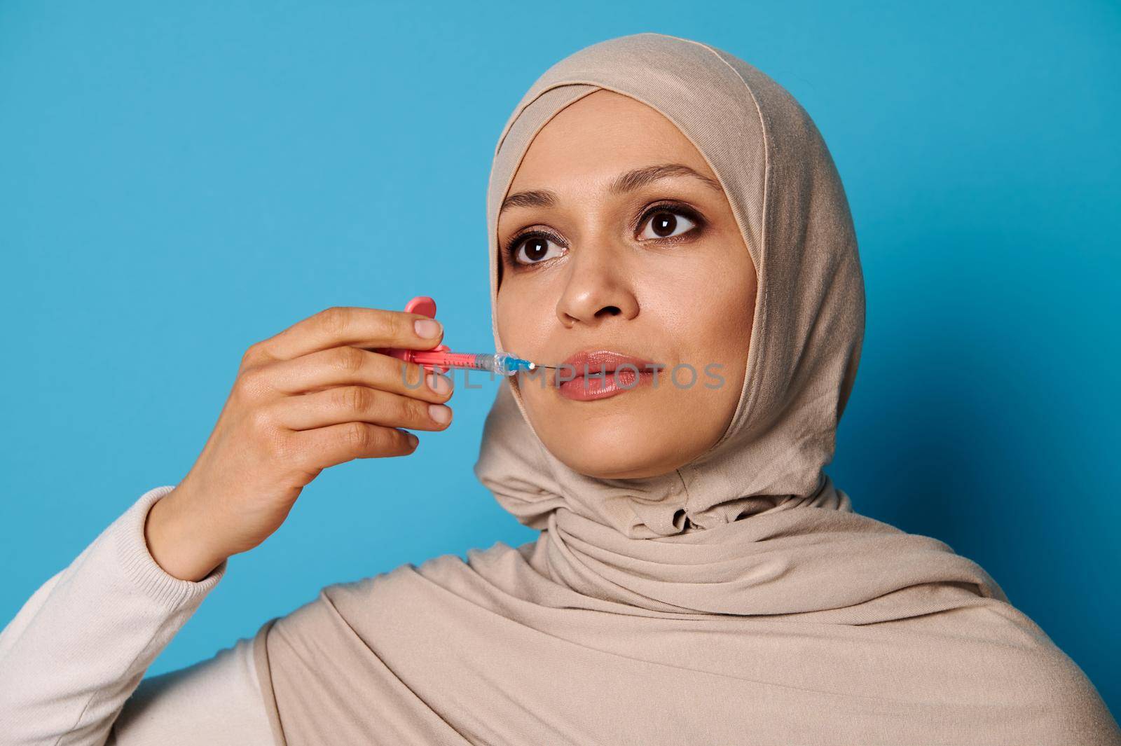 Closeup of beautiful Muslim woman in hijab holding a syringe with beauty injection near her lips. Lips augmentation concept in injection cosmetology. Copy space, blue background