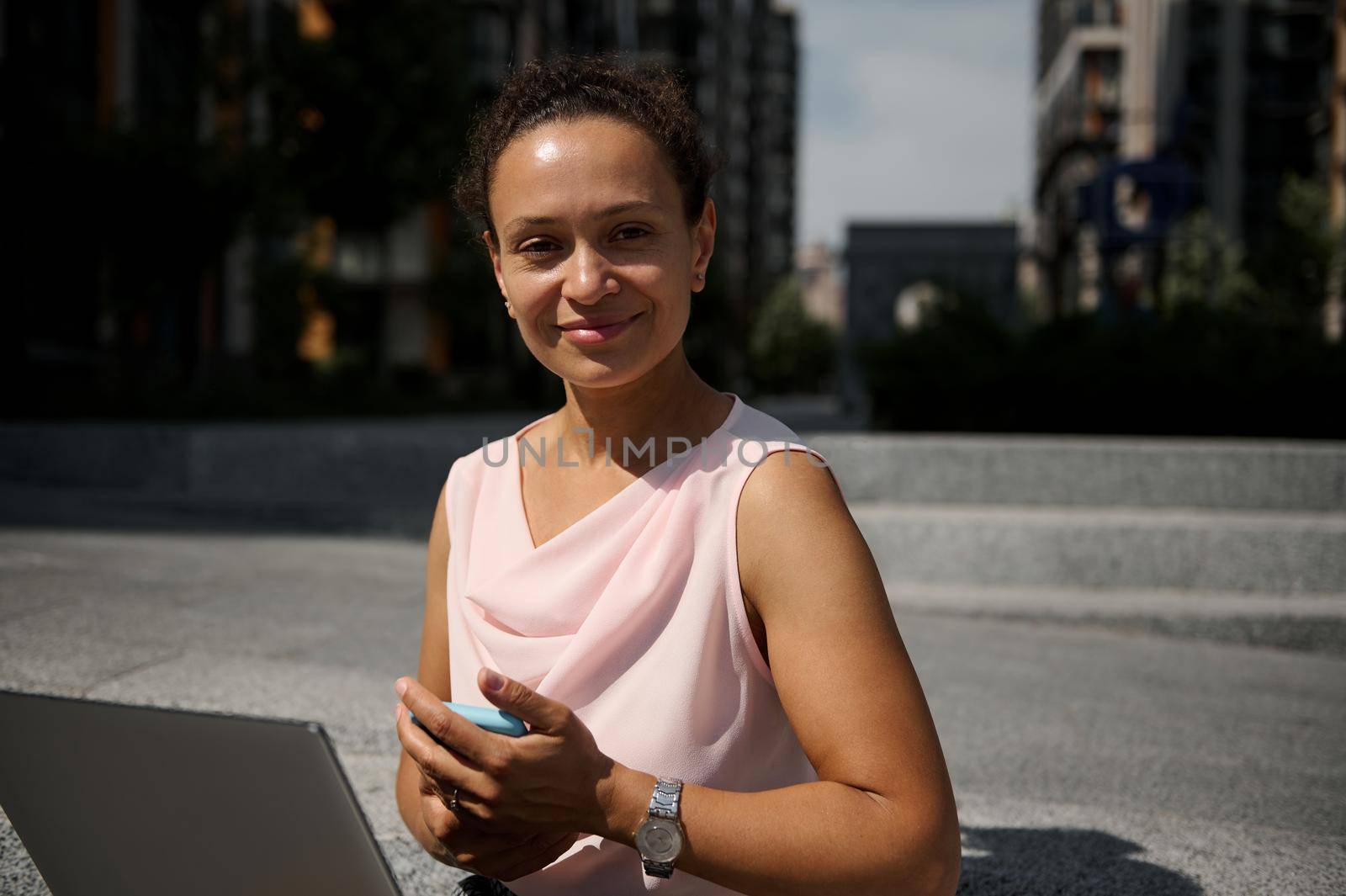 Confident close-up portrait of a beautiful mixed race business woman with laptop and smartphone looking at the camera sitting on steps on the background of urban tall buildings