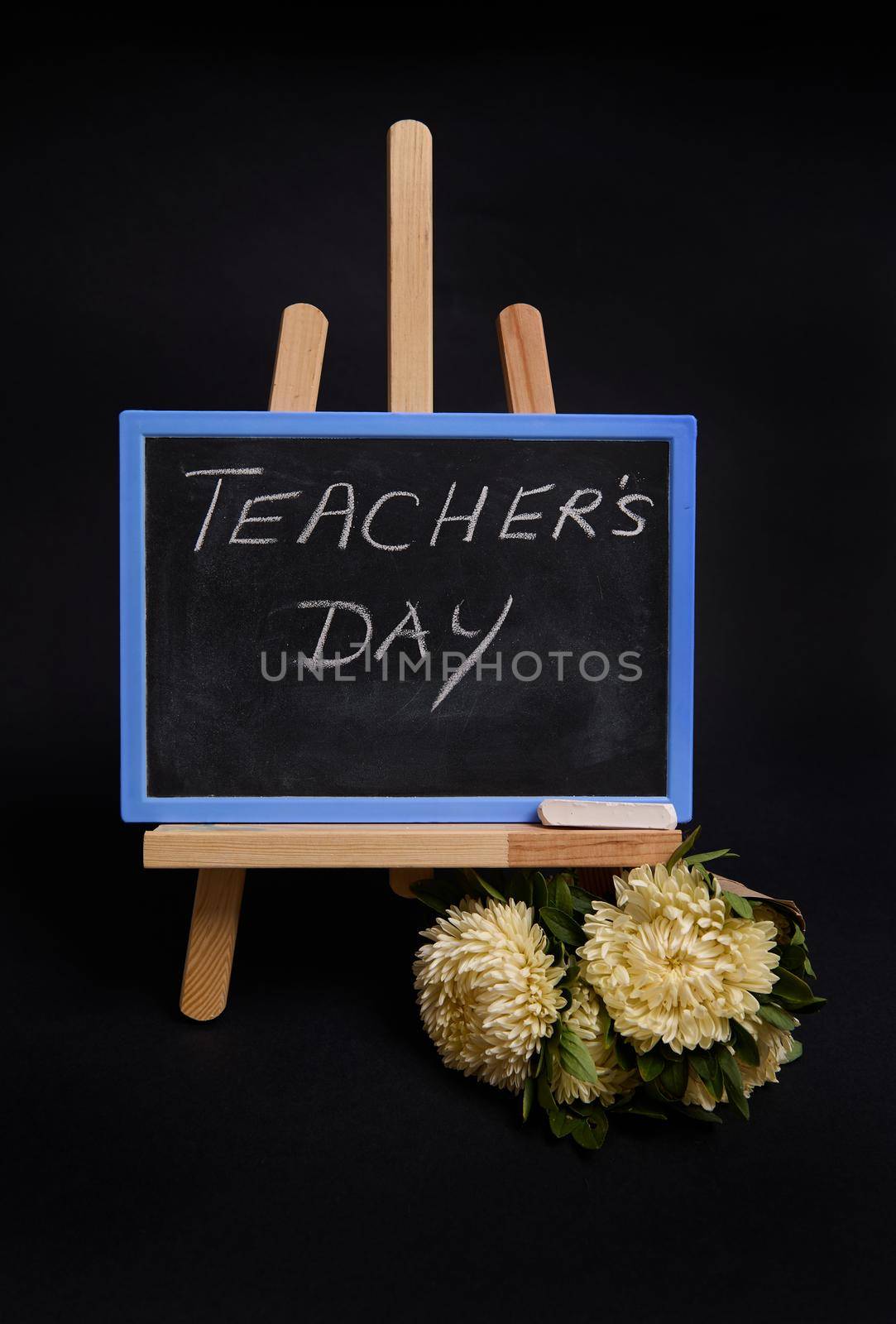 Close-up of a chalk blackboard with lettering Teacher's Day, standing on a wooden table easel, next to black alarm clock, isolated on black background with copy space. by artgf