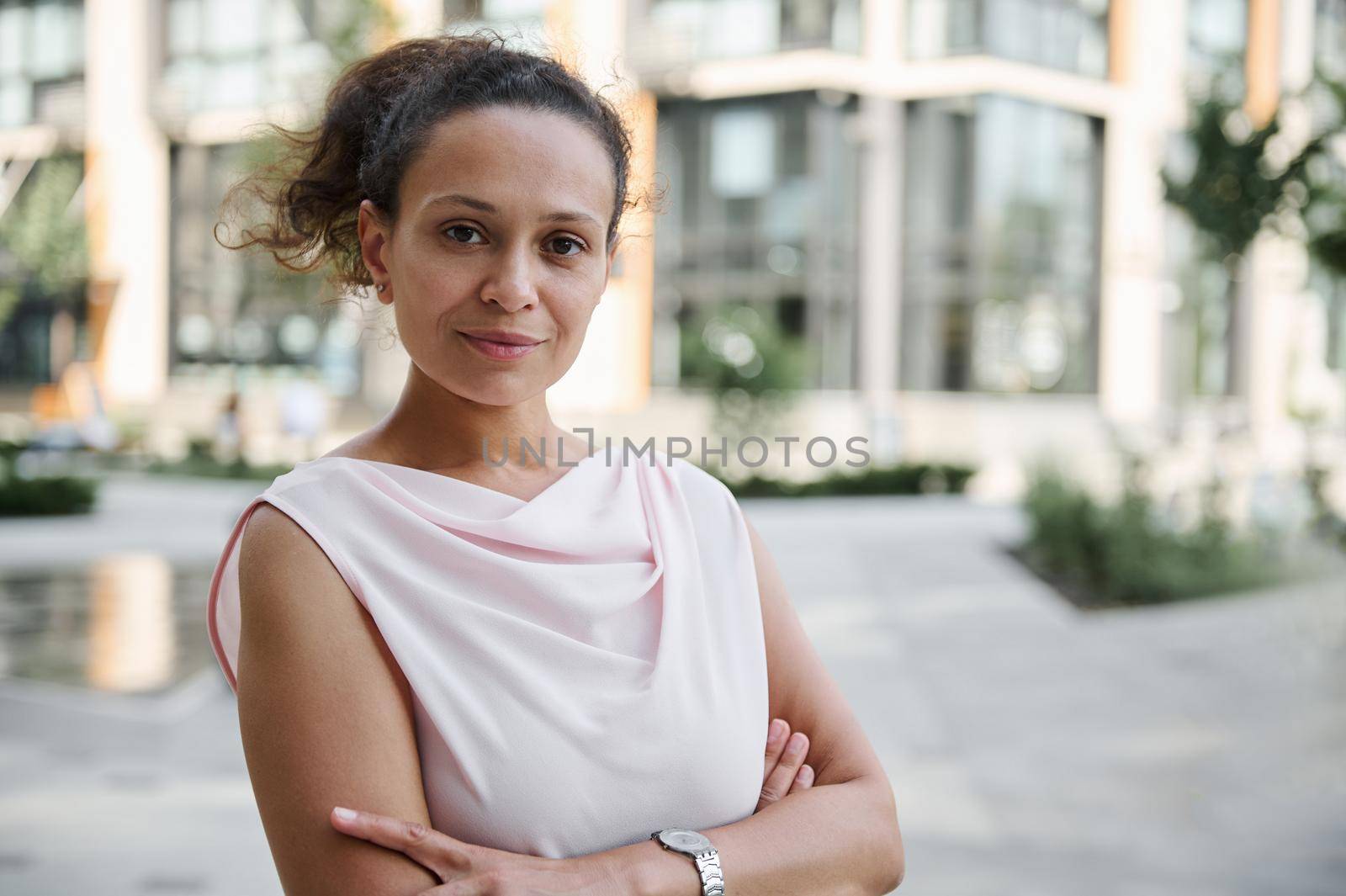 Close-up confident portrait of a successful middle aged business woman in casual attire posing to camera with crossed rams on the background of high buildings. Urban city backdrop