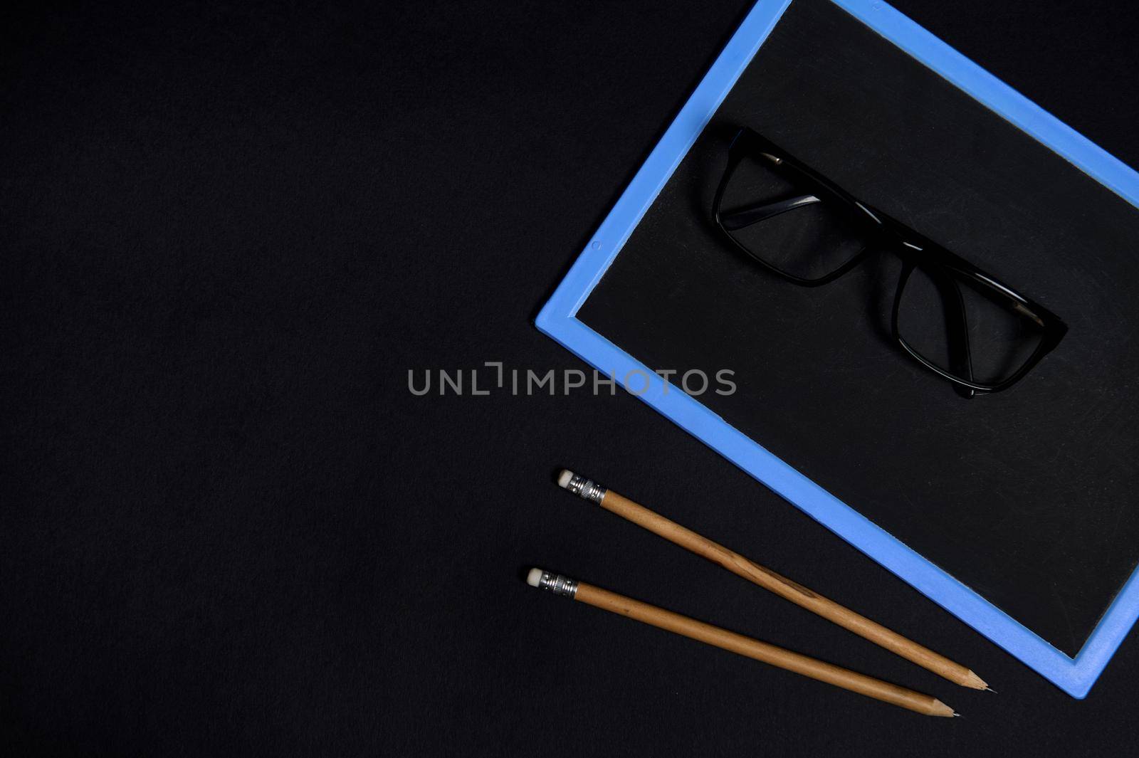 Flat lay of eyeglasses on an empty blank chalkboars and two wooden pencils on black surface background with coy space