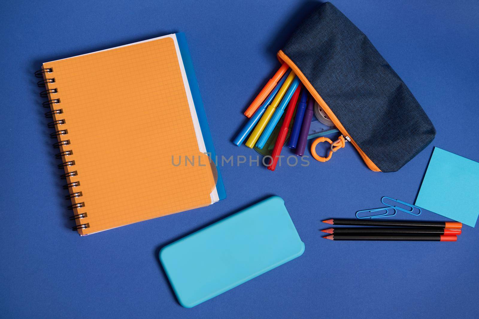 Top view of school office supplies and a smartphone lying screen down on a blue background . Flat lay composition of stationery in two contrasting colors, blue and orange with copy space