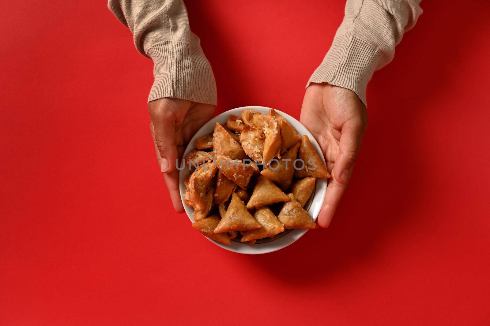 Hands holding a delicious and sweet plate full of fresh traditional Moroccan handmade sweets, isolated on red background. Space for text. Arabic traditional oriental sweets on the festive table