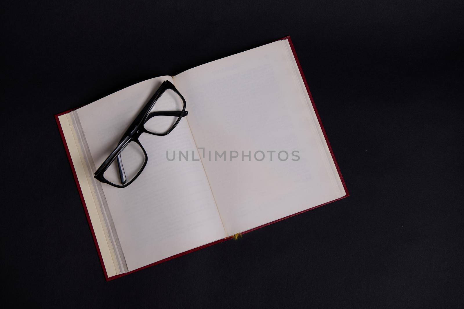 Flat lay composition of eyeglasses on an opened book in hard red cover, isolated over black background with space for text. Teacher's Day concept, Knowledge, literature ,reading, erudition