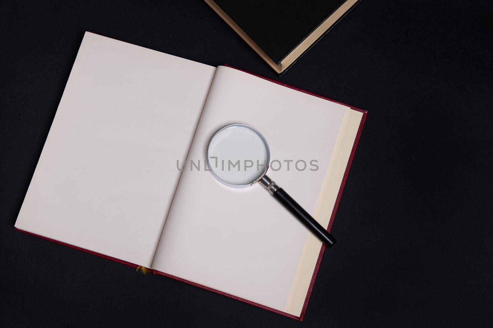 Flat lay composition of a magnifying glass on an opened book in hard red cover, isolated over black background with space for text. Teacher's Day concept, Knowledge, literature ,reading, erudition by artgf