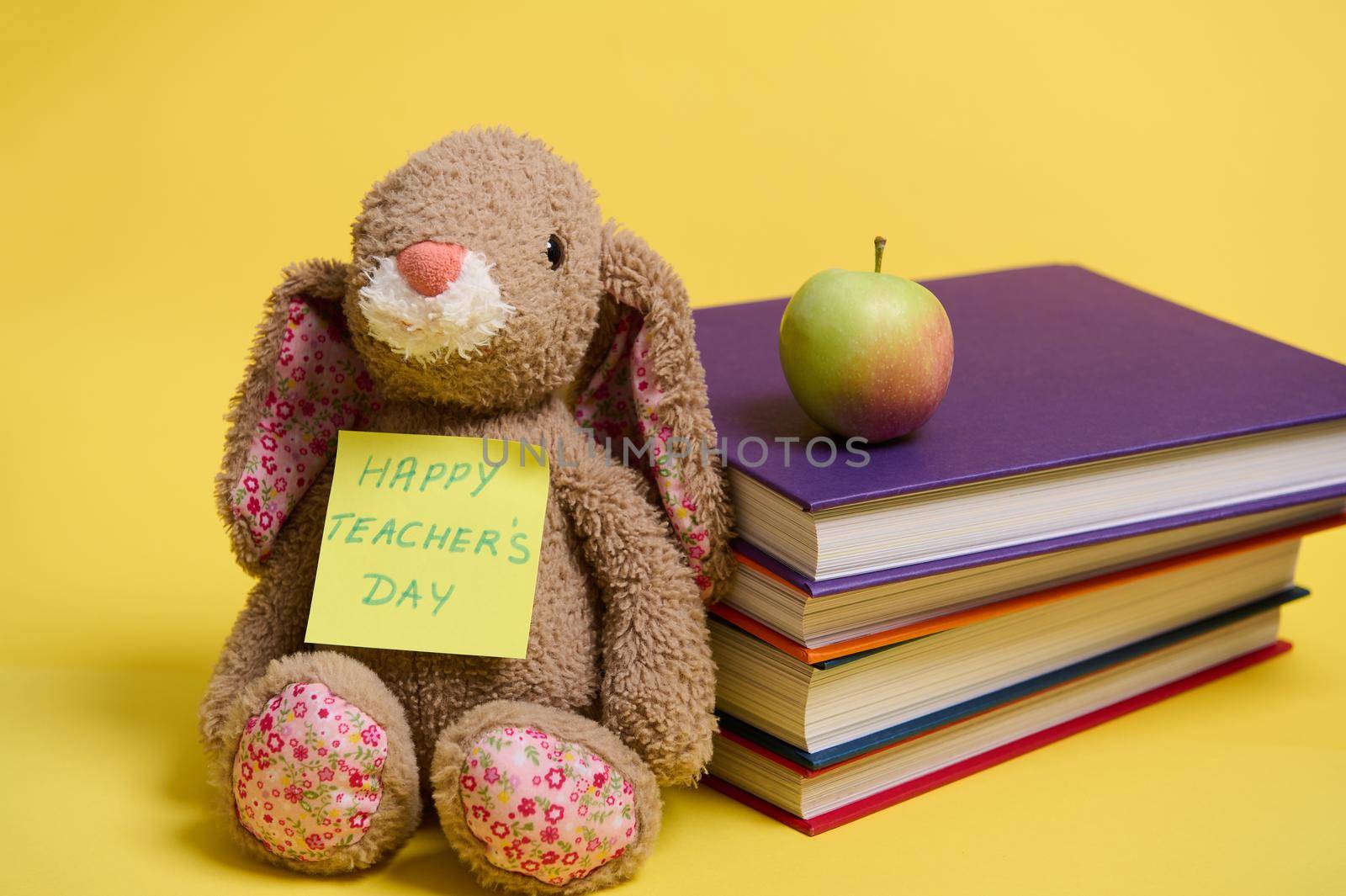 Close-up of plush toy rabbit with lettering Happy Teacher's Day on yellow note paper, leaned on a stoke of colorful books, yellow background with space for text