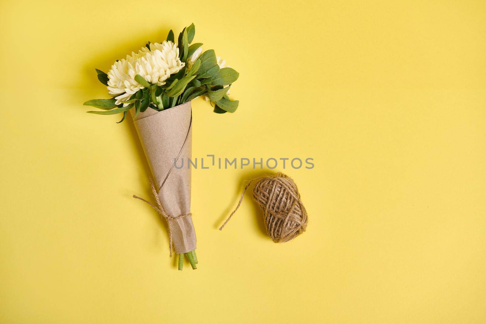 Flat lay floral arrangement of a delicate beautiful bouquet of autumn asters flowers in kraft wrapping paper tied with a rope and a skein of rope on a yellow background with space for text.