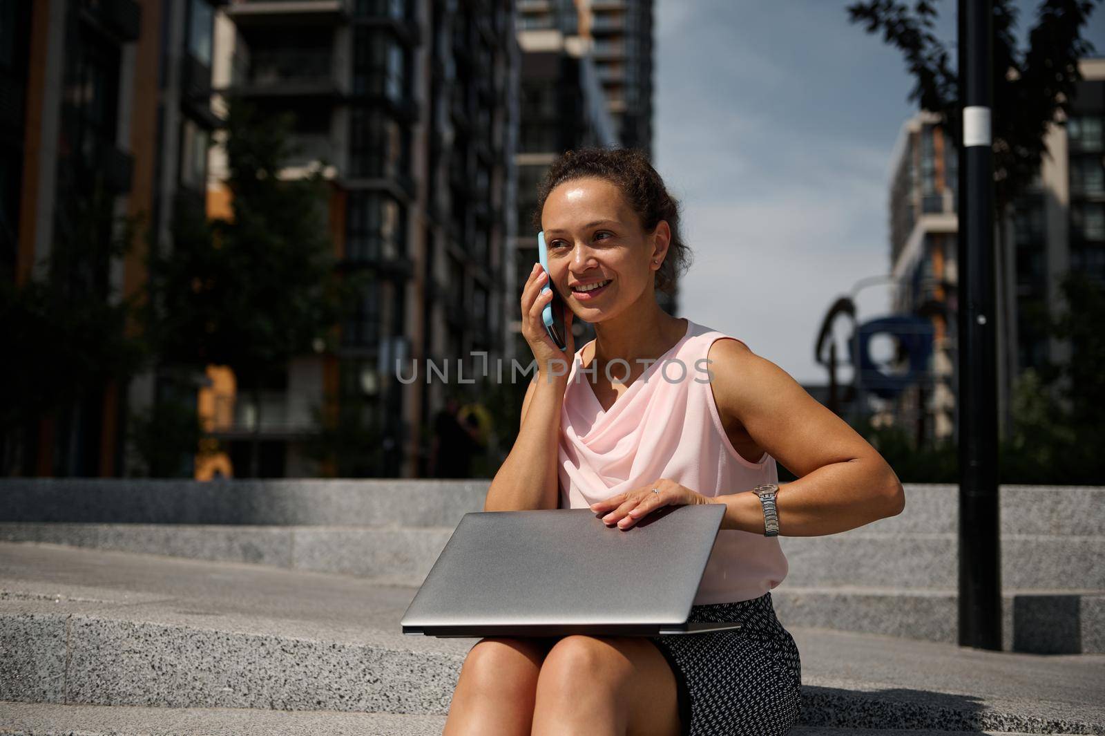 Adorable mixed race African American woman talking on mobile phone, sitting on the steps with a laptop on the urban high buildings background. Business woman at lunch break time