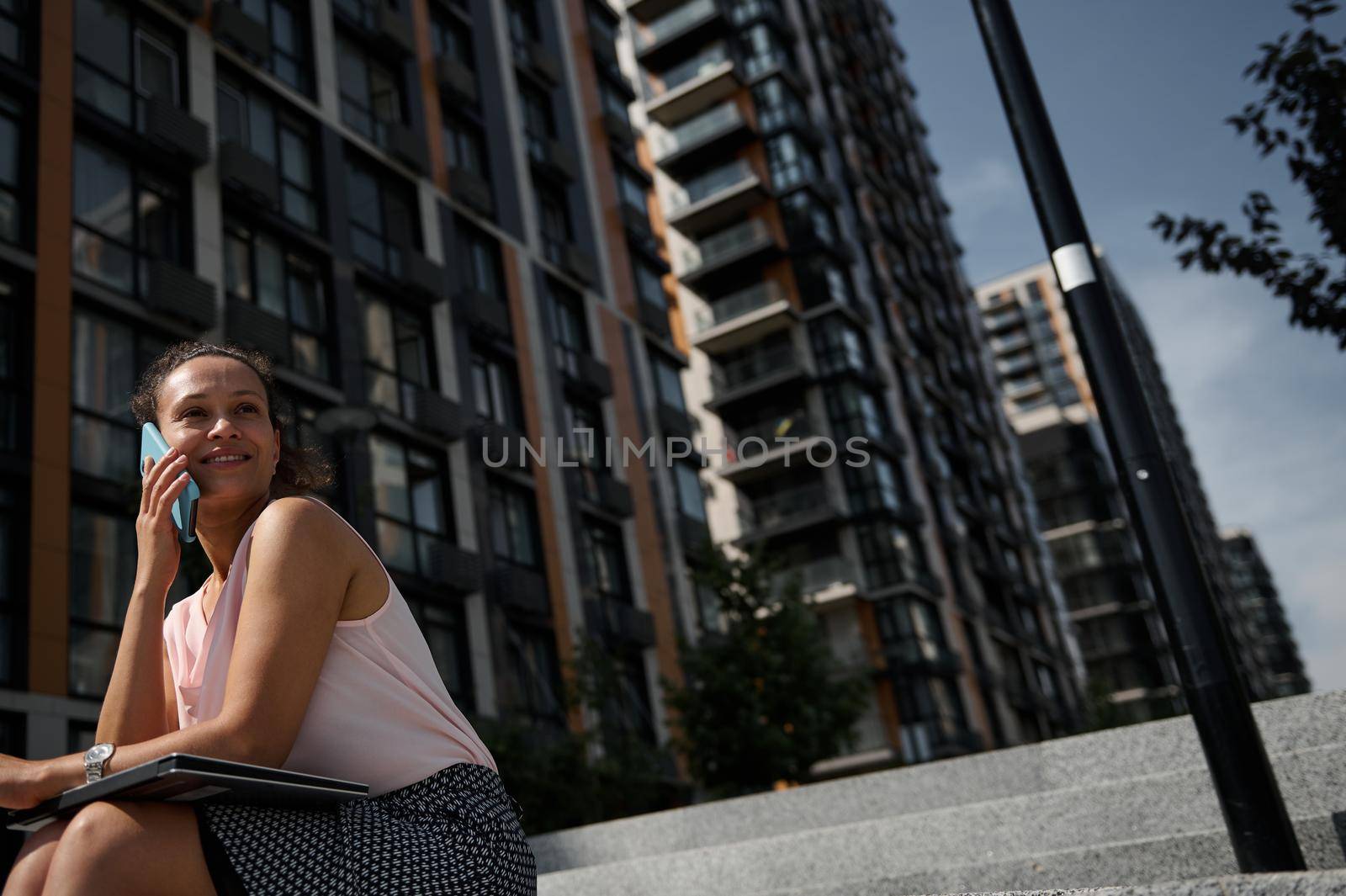Portrait of a beautiful African American woman talking on mobile phone, looking away, posing to camera on urban high buildings background. Business, office, freelance, remote work, online concept