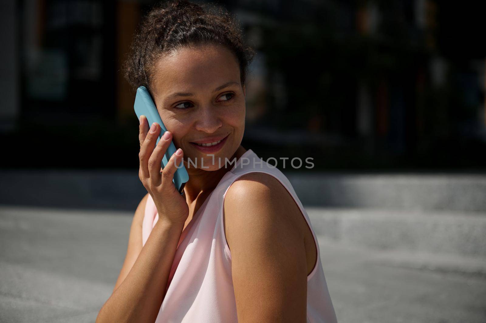 Close-up portrait of a beautiful African American woman talking on mobile phone, looking away, posing to camera on urban buildings background by artgf