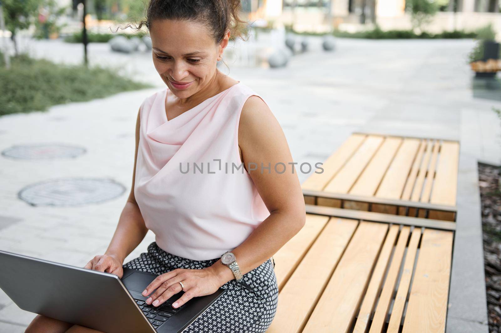 Beautiful young African American woman freelancer working on laptop while sitting on wooden bench in city