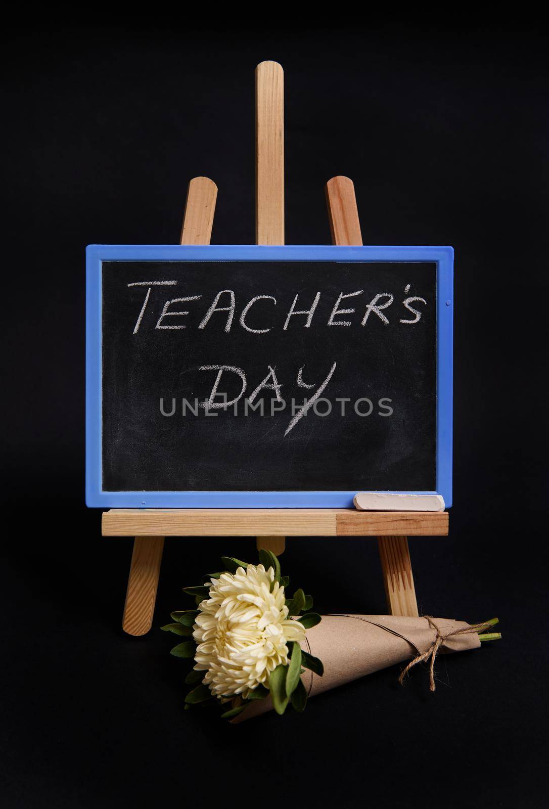 Close-up of a bouquet of asters flowers next to a chalk chalkboard with lettering Teachers Day, standing on a wooden table easel, isolated on black background with copy space. by artgf