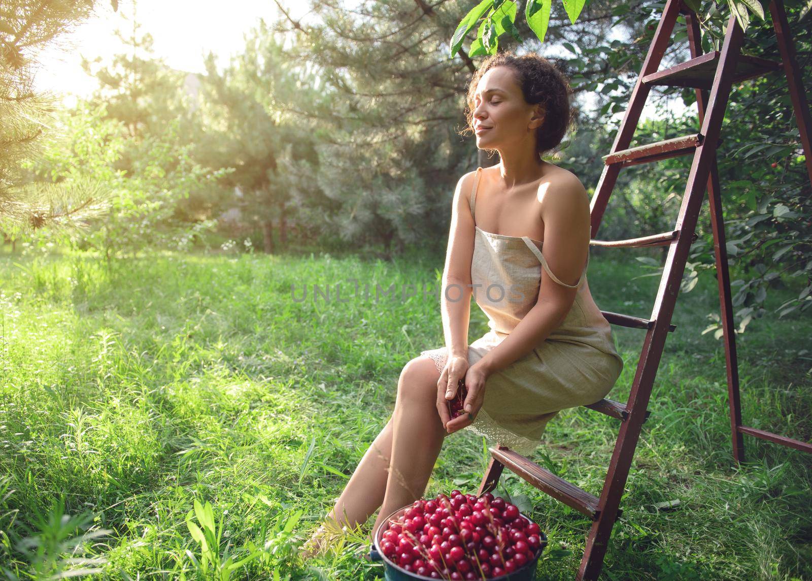 Beautiful woman in a linen dress sitting on a ladder next to a bucket of cherries in the orchard at sunset. Beautiful sun rays fall into the garden on a summer day by artgf