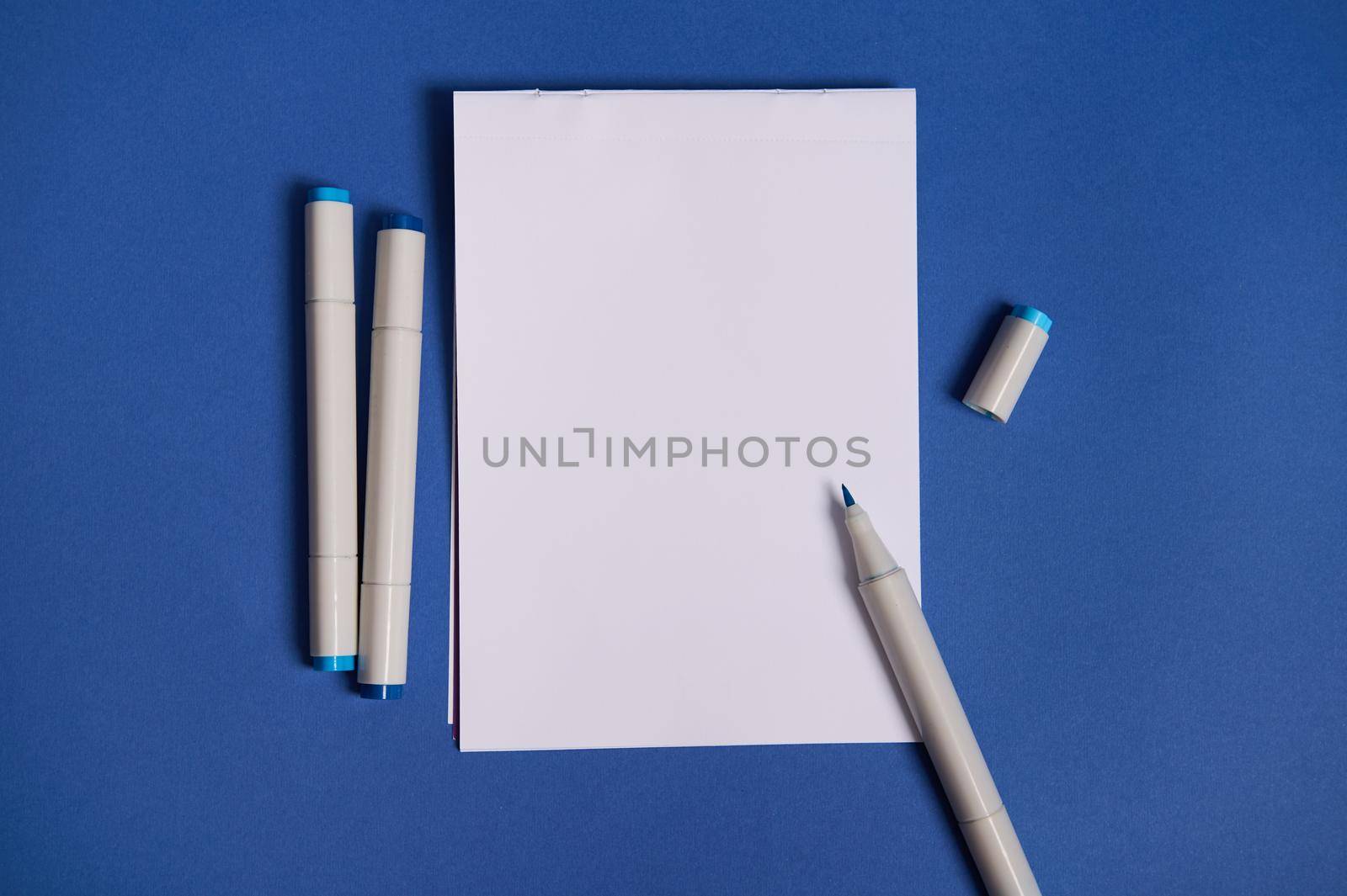 Dichromatic flat lay composition with watercolor markers or felt-tip pens with a white blank empty paper sheet with copy space, isolated on blue background. by artgf