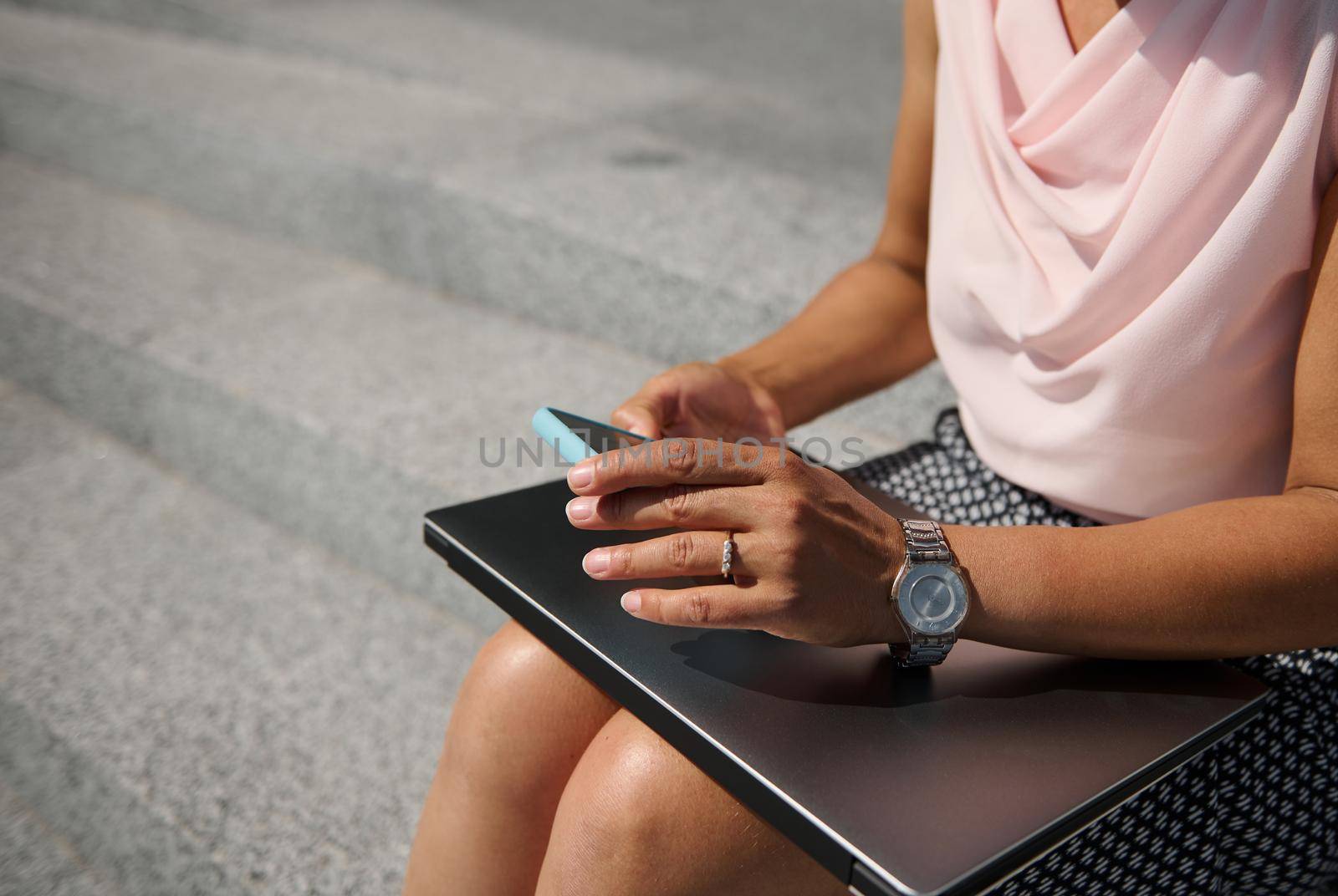 Close-up, cropped view with soft focus on business woman hands with stylish hand watch, holding a smartphone , sitting on steps with a laptop. Business, freelance, online and distant work concept by artgf