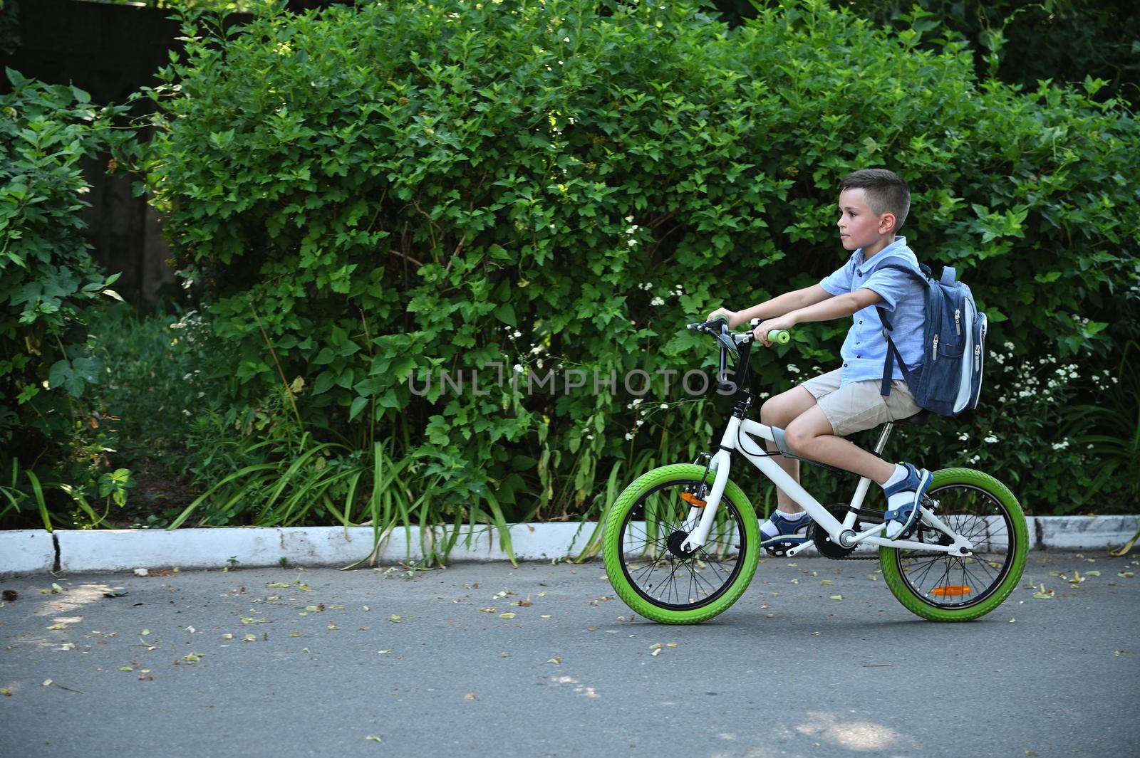 Side full-length portrait of school boy on a bicycle at asphalt road in early morning. Child on bike in the city