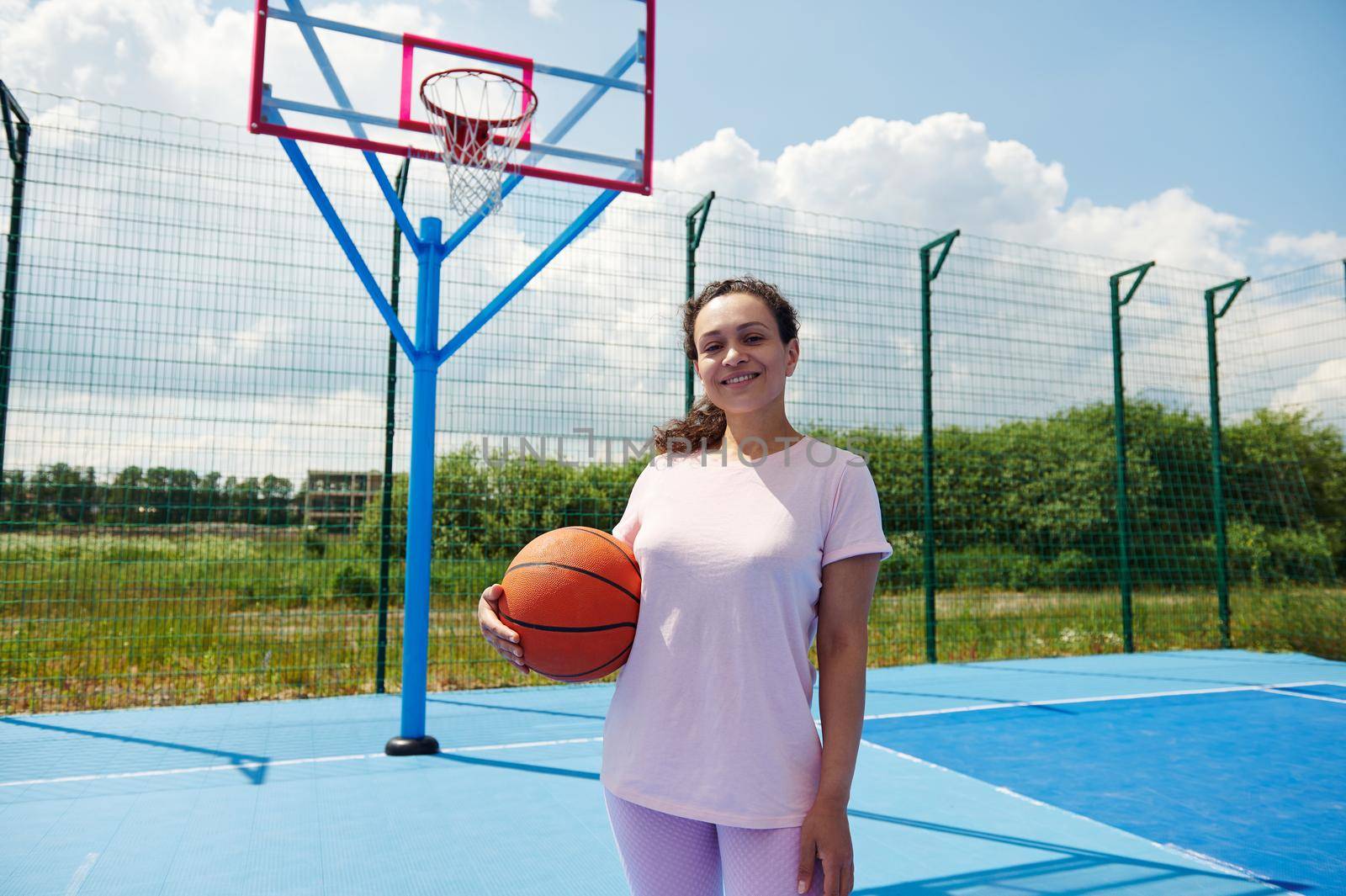 African American female basket ball player standing on a court and smiles holding a basketball under arms by artgf