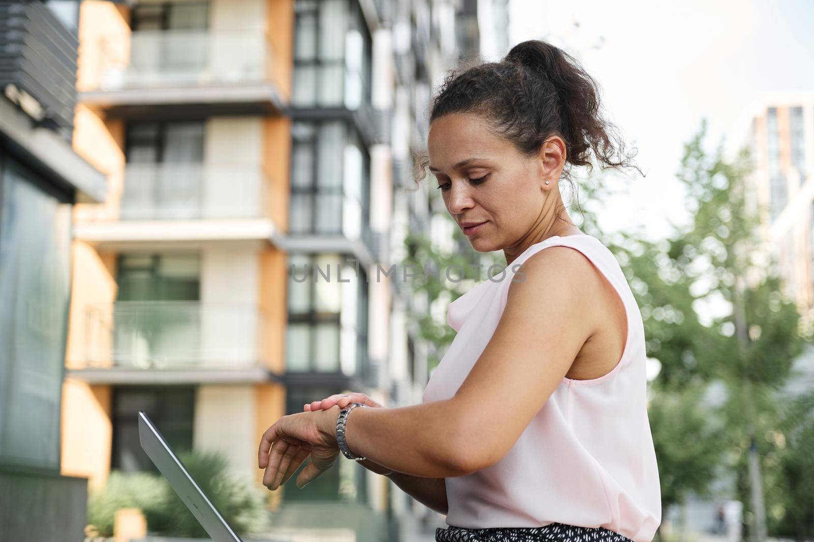 Busy freelancer woman checking time on hands watch while resting in city, sitting on a street wooden bench during coffee break time by artgf